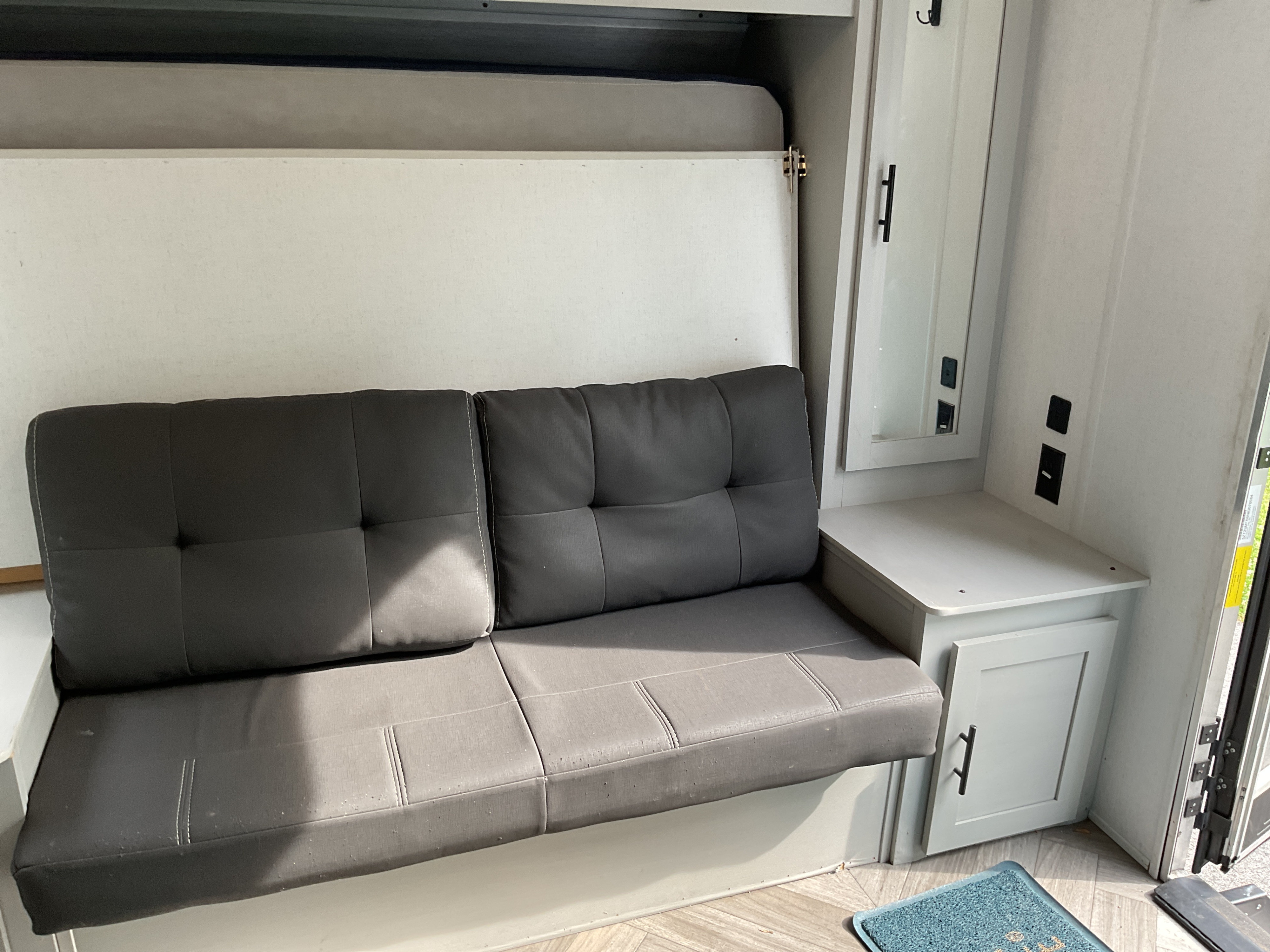 This RV rental interior features a gray convertible couch, a neatly folded Murphy bed above, and built-in storage with a mirror and outlets, maximizing compact living space.. Forest River Salem 2021