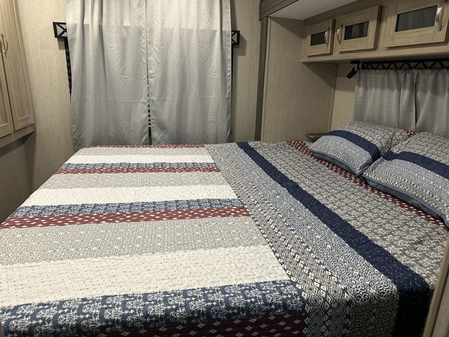 An RV rental bedroom with a bed, colorful patterned quilt, pillows, window curtains, and overhead cabinets.. Coachmen Freedom Express 259FKDS 2021