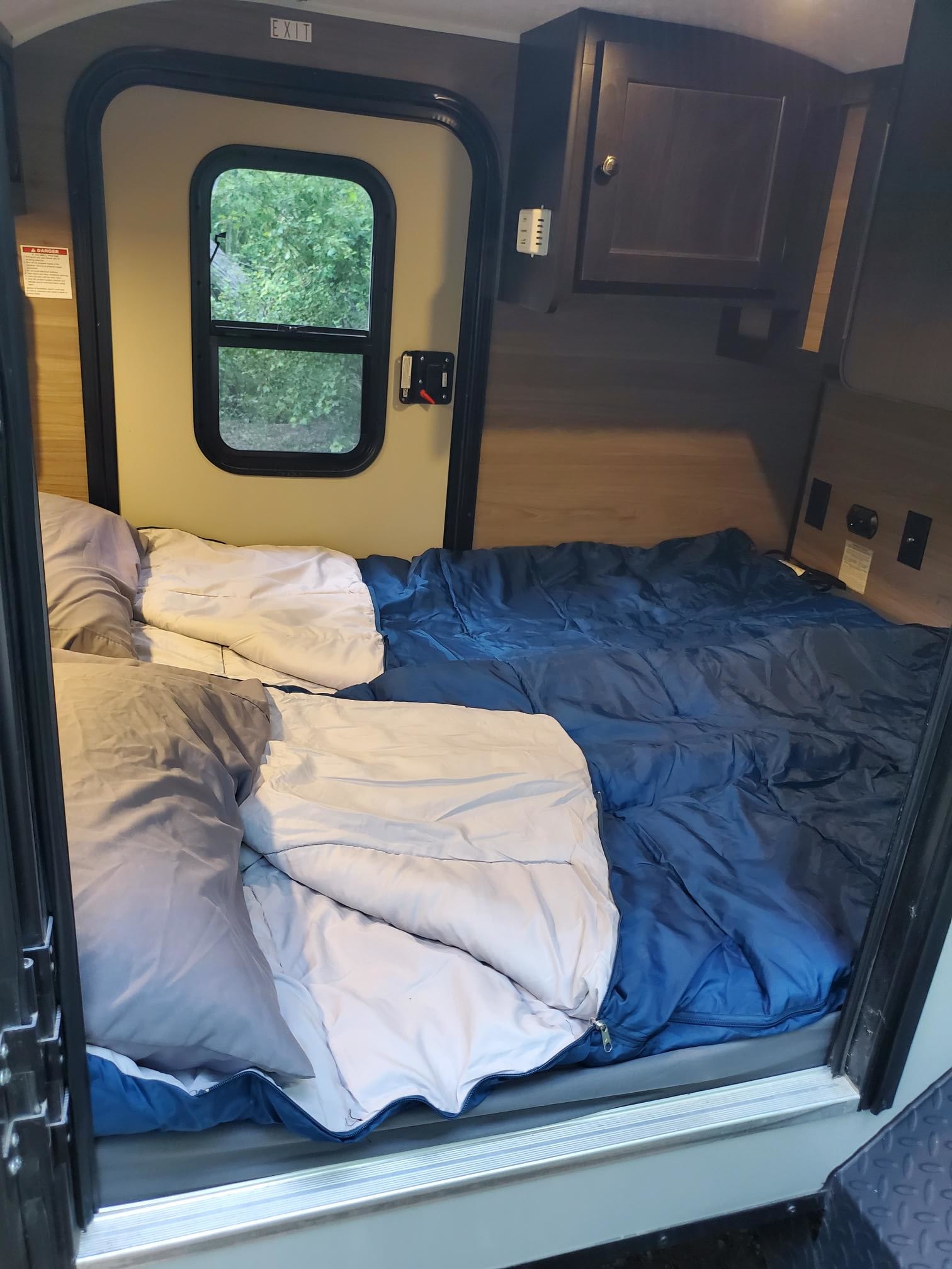 Inside a compact rv rental: a cozy bed with sleeping bags, pillows, and an exit door leading to nature. Ready for travel.. Braxton Creek Bushwhacker 2024