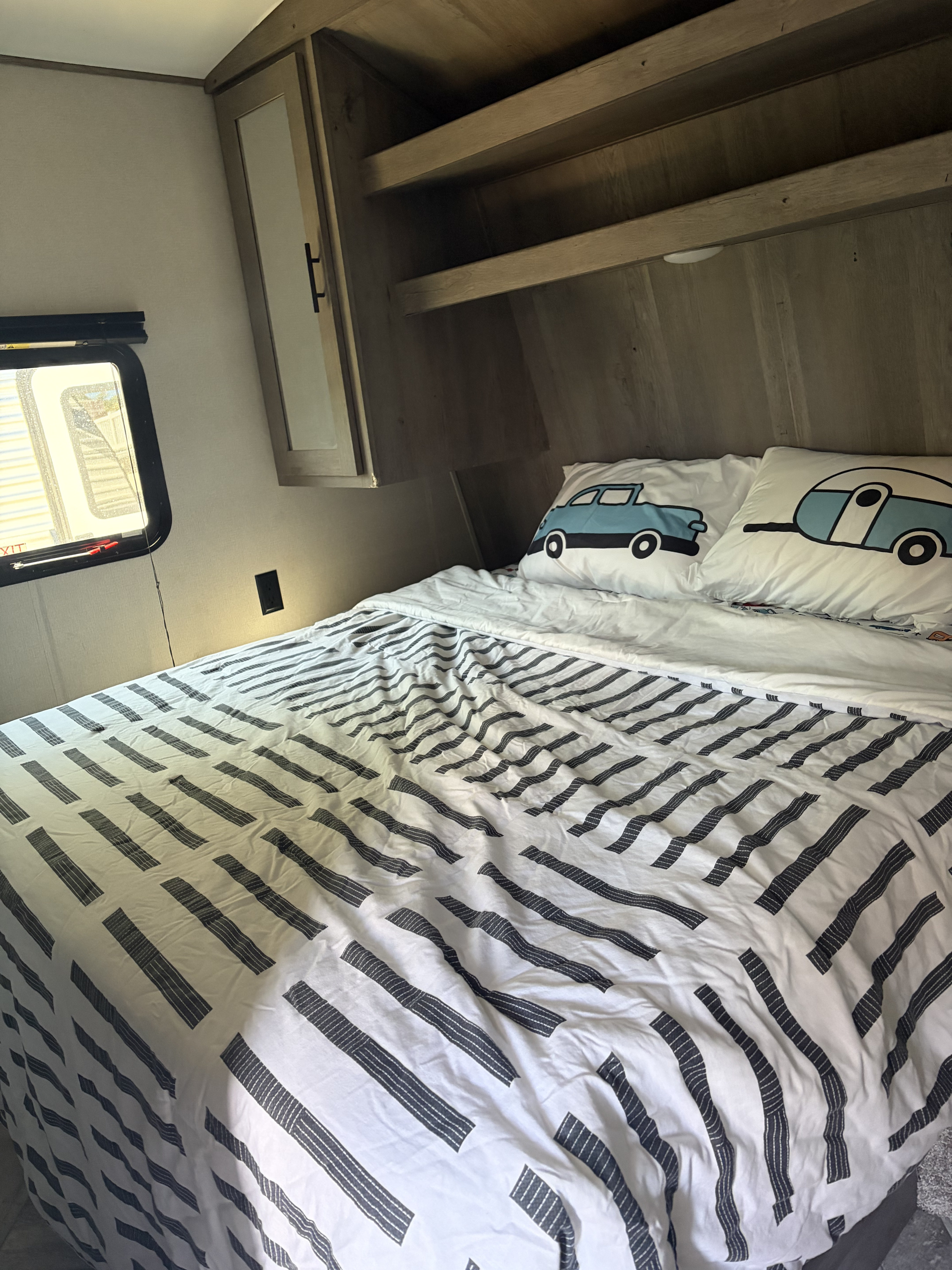Cozy RV rental bedroom features striped bedding, fun car & trailer pillows, window, and wood storage cabinets.. Keystone RV Springdale 2021