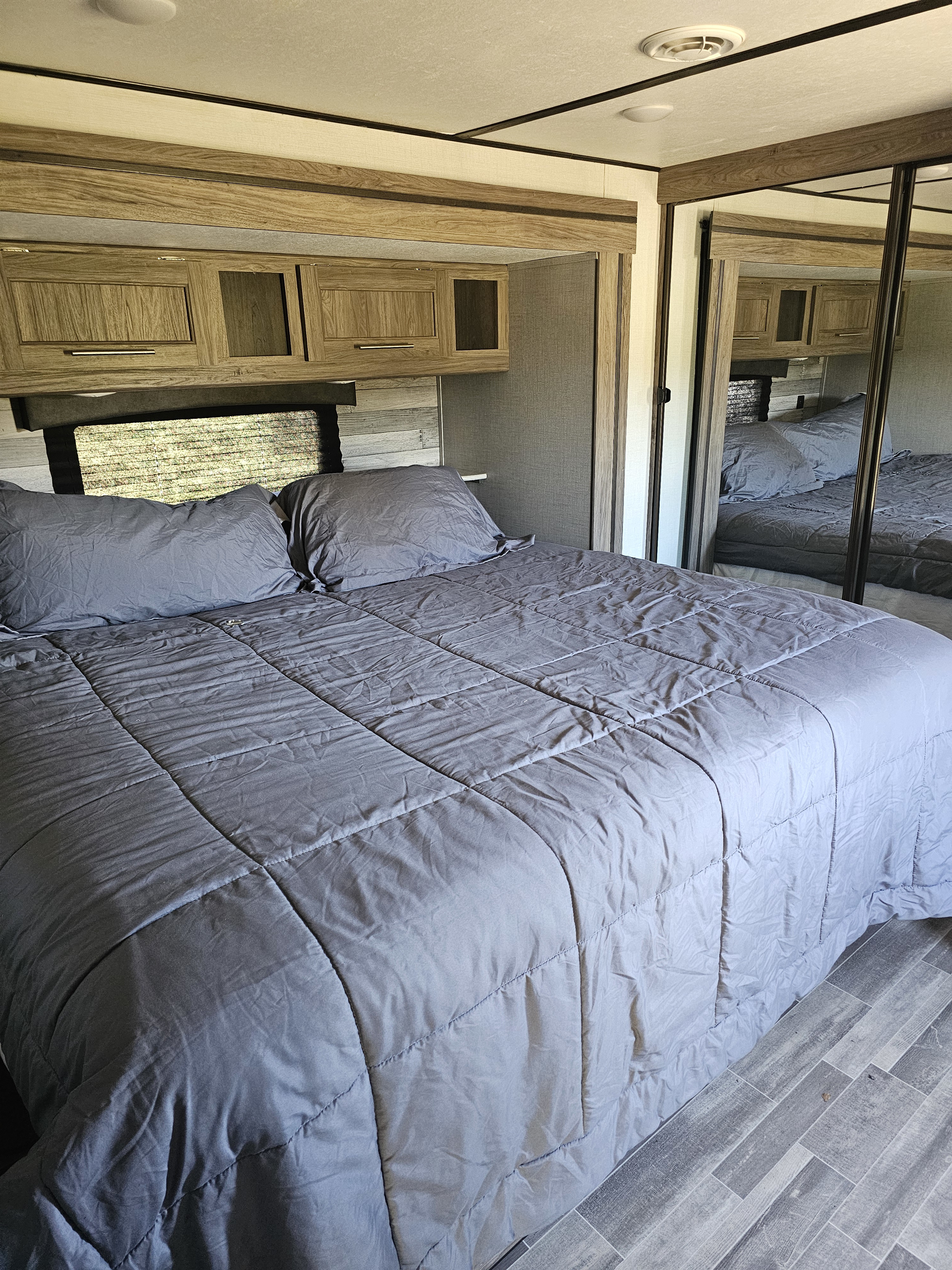 Spacious RV rental bedroom featuring a bed with grey bedding, wood-look overhead cabinets, and a mirrored closet.. KZ Sportsman 2022