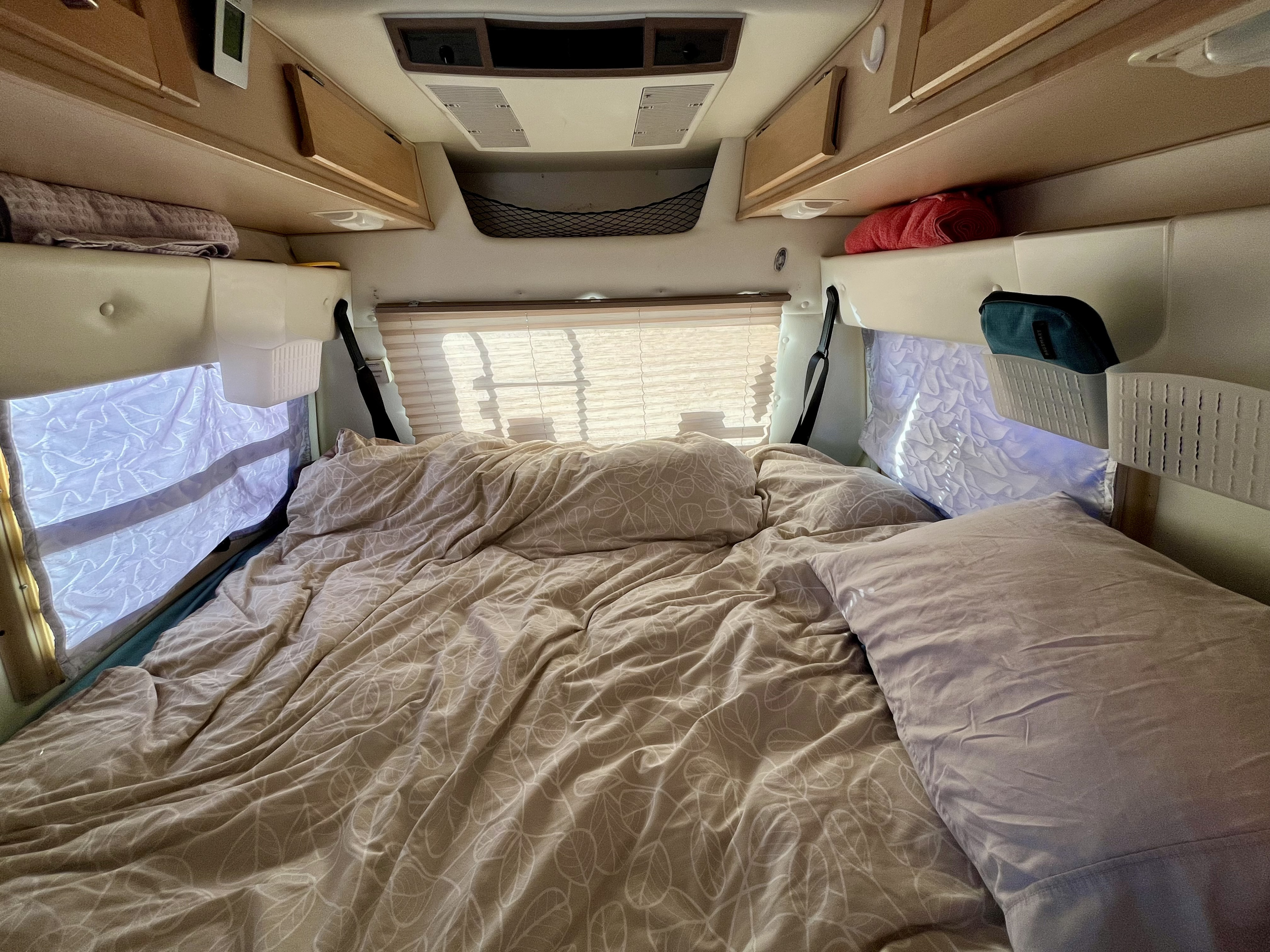 Cozy bed in an RV rental, featuring light bedding with leaf patterns, window blinds, and overhead storage for travel essentials.. Dodge Leisure Travel Wide Body 2002
