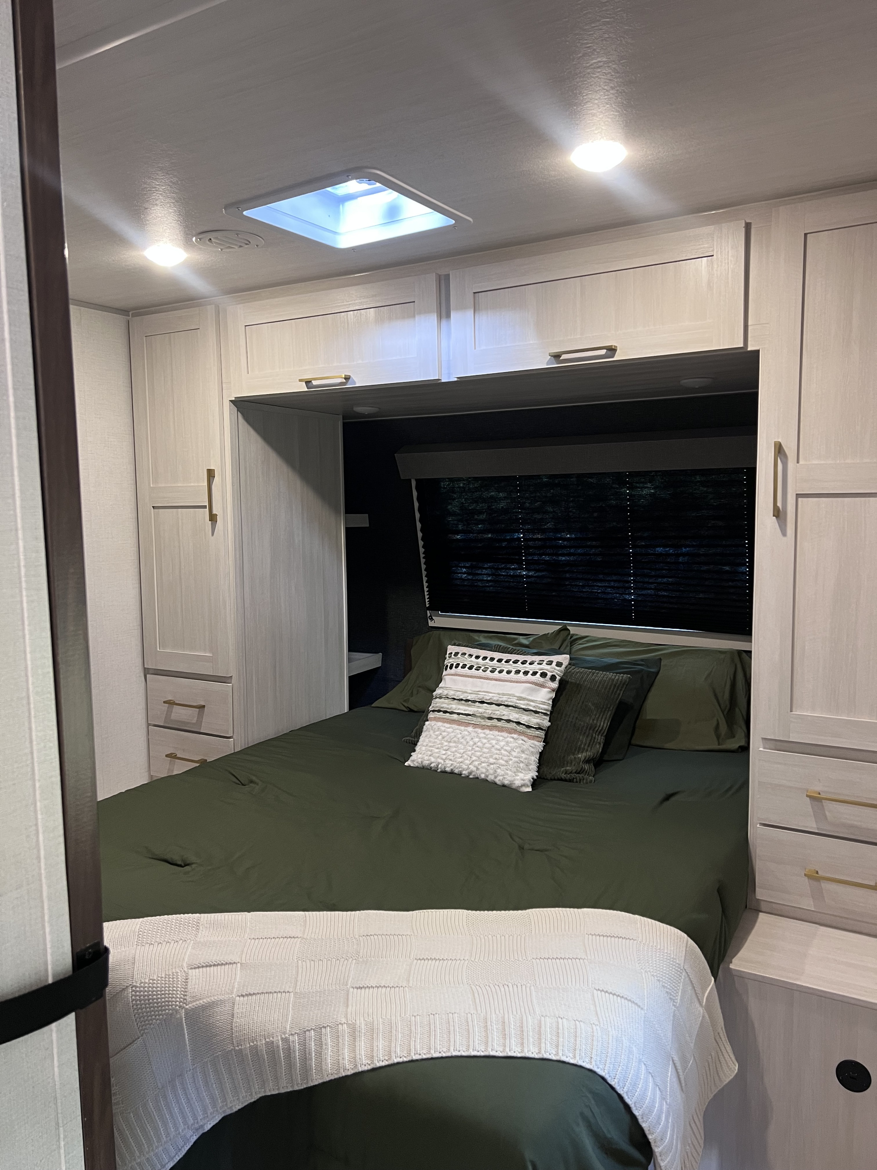 Cozy RV rental bedroom featuring a bed with dark green bedding, a white knit blanket, and light wood cabinets.. Forest River Surveyor Legend 2026