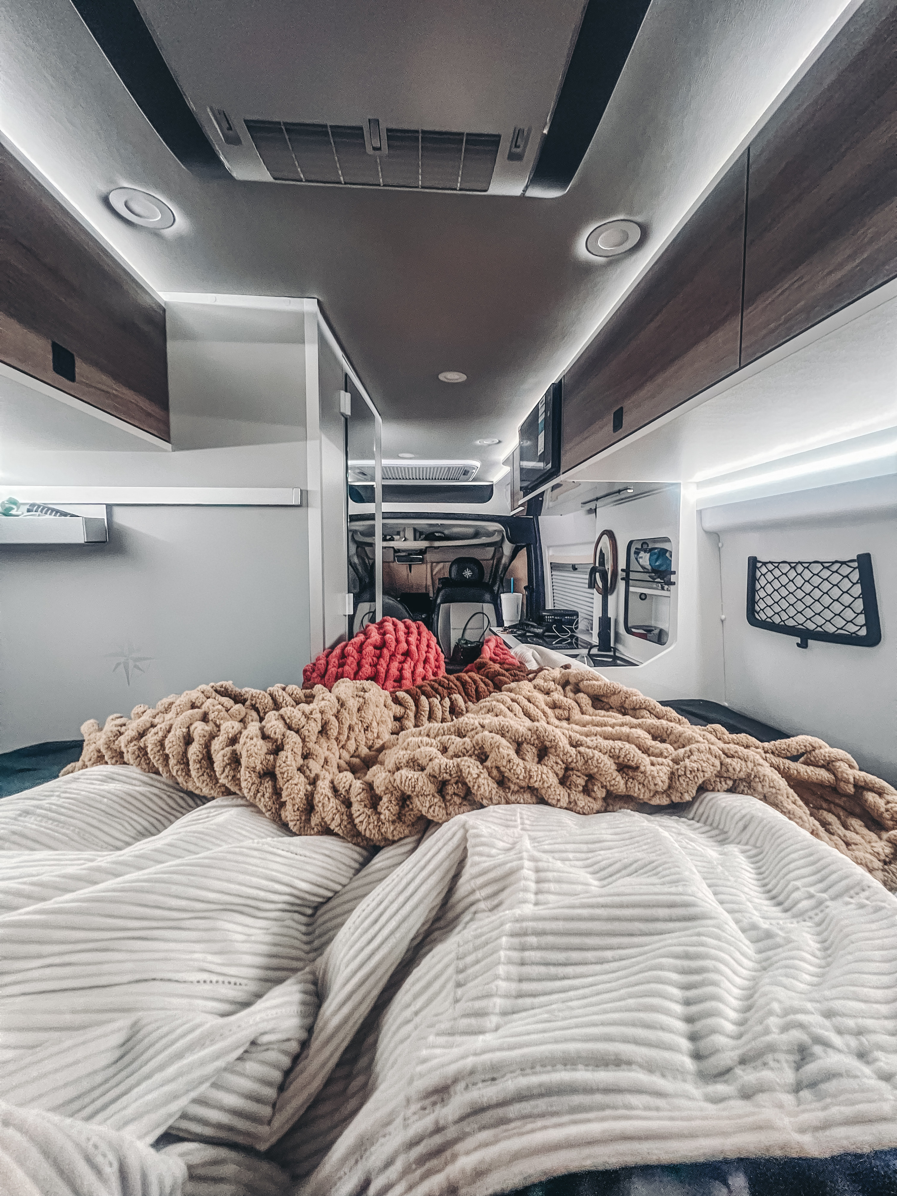 Cozy RV rental interior from the bed, featuring chunky blankets, overhead lights, and a view to the cockpit/kitchen.. Roadtrek/Westfalia Wave SRT 2026