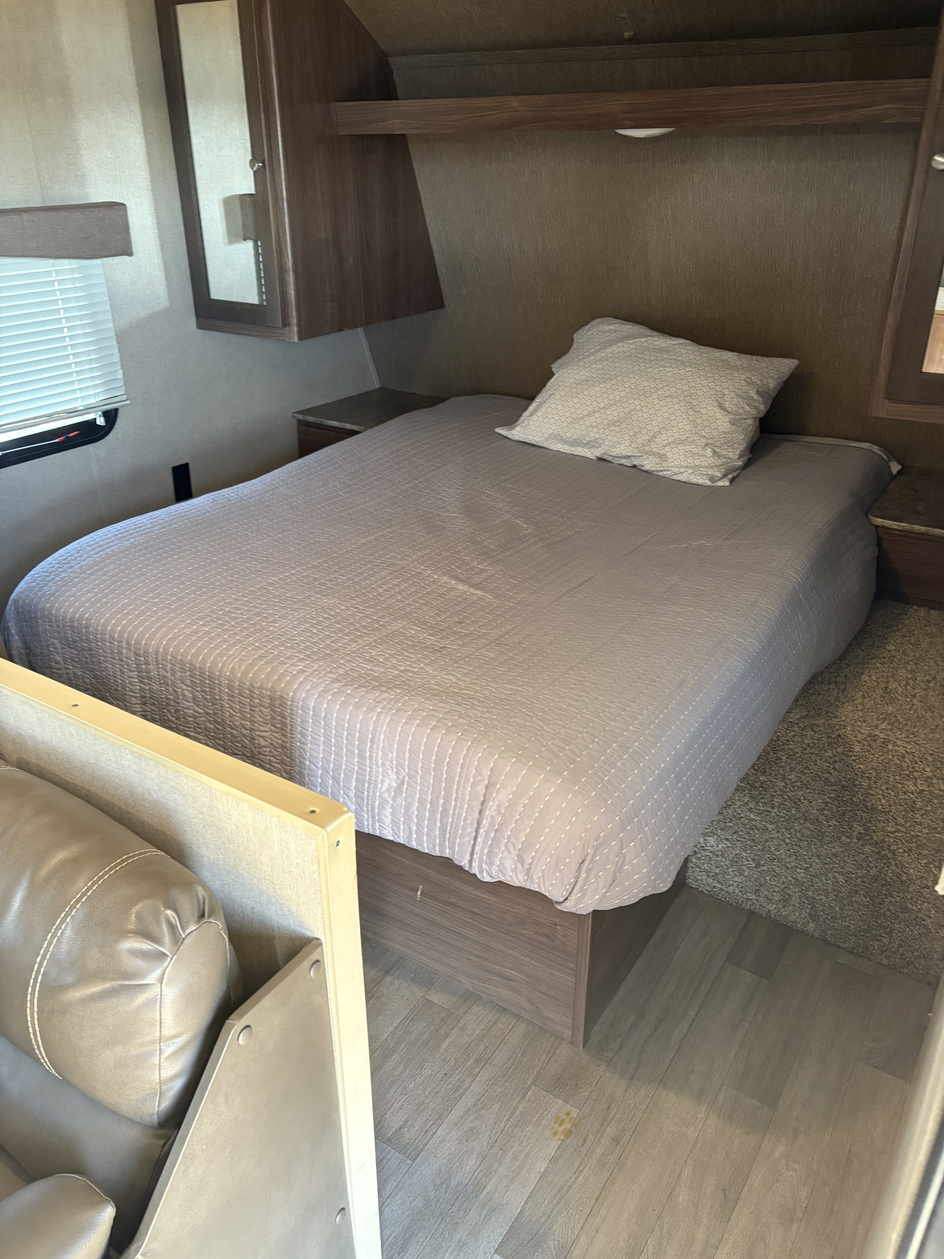 RV rental bedroom view featuring a neatly made bed with a grey quilted comforter, wooden overhead cabinets, and a window with blinds.. Coleman Lantern 2018