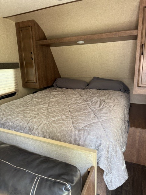 RV rental bedroom with a comfortable bed, gray bedding, overhead storage, and a partial view of a sofa.. Starcraft Autumn Ridge Outfitter 2018