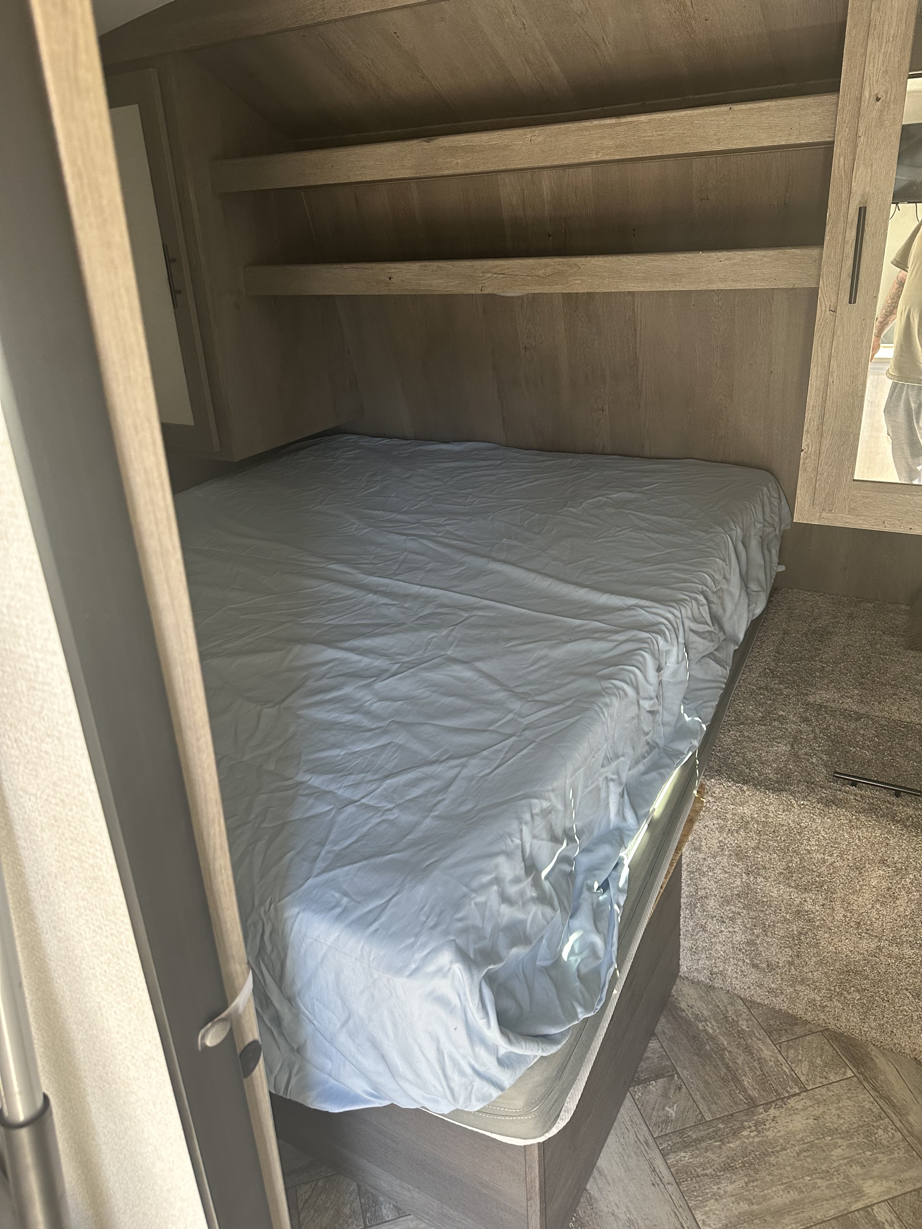 Compact rv rental sleeping area featuring a mattress with light blue sheets, wooden shelves, and mixed carpet/wood flooring.. Keystone RV Springdale 2022
