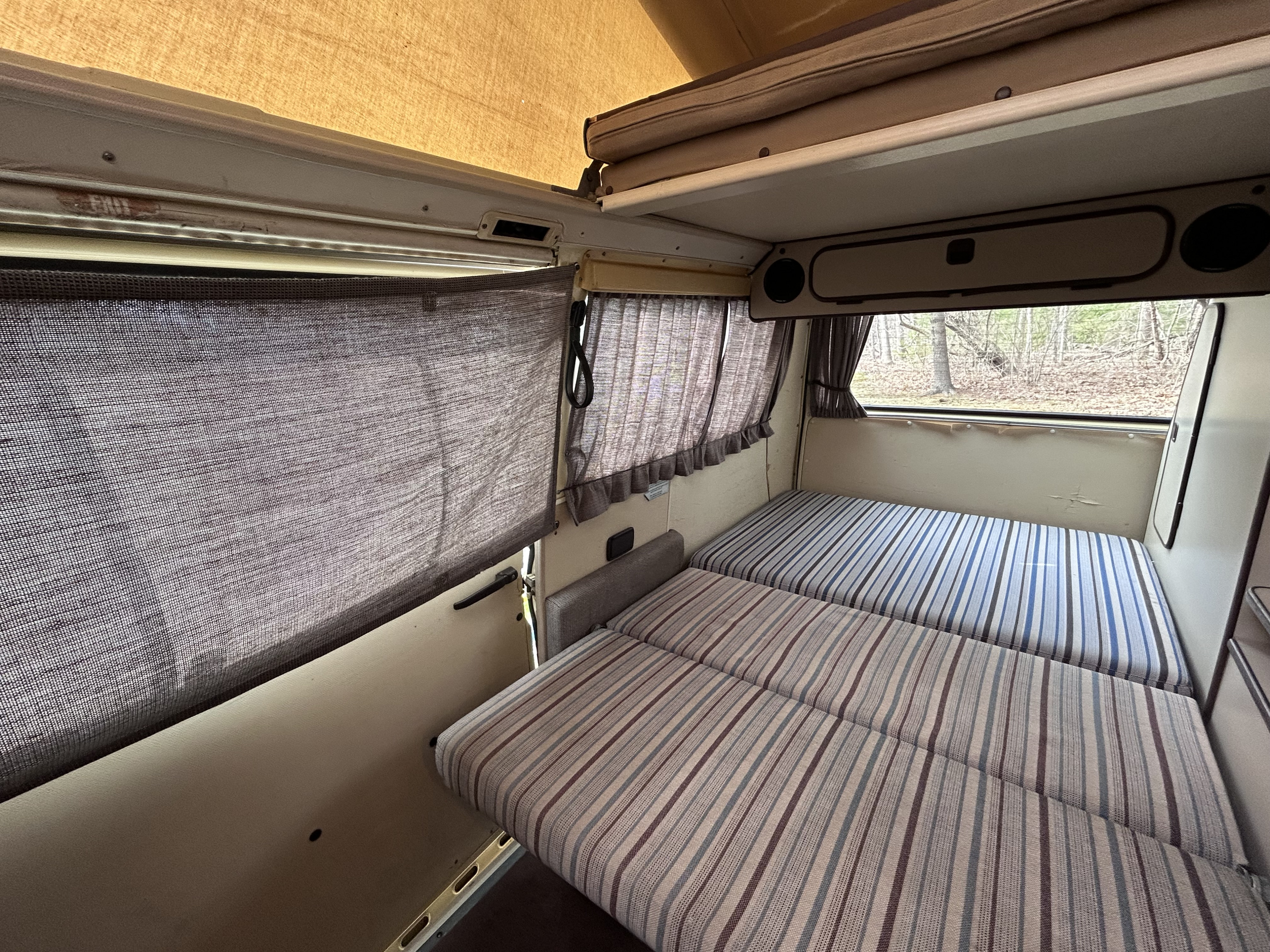 Cozy camper van interior from an RV rental, featuring striped beds, a pop-top, and window privacy screens for comfortable travel.. Volkswagen T3 Westfalia 1984