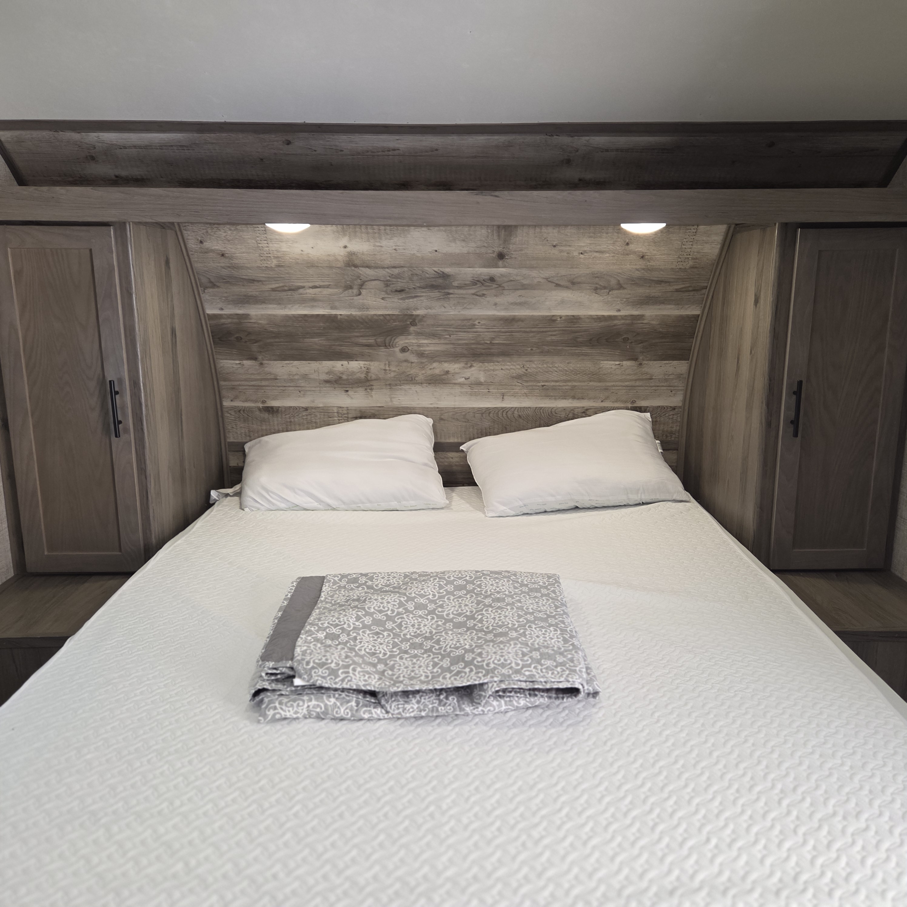 An RV rental bedroom with a neatly made bed, white pillows, and a gray patterned blanket. Rustic wood paneling and cabinetry.. Gulf Stream Enlighten 2021