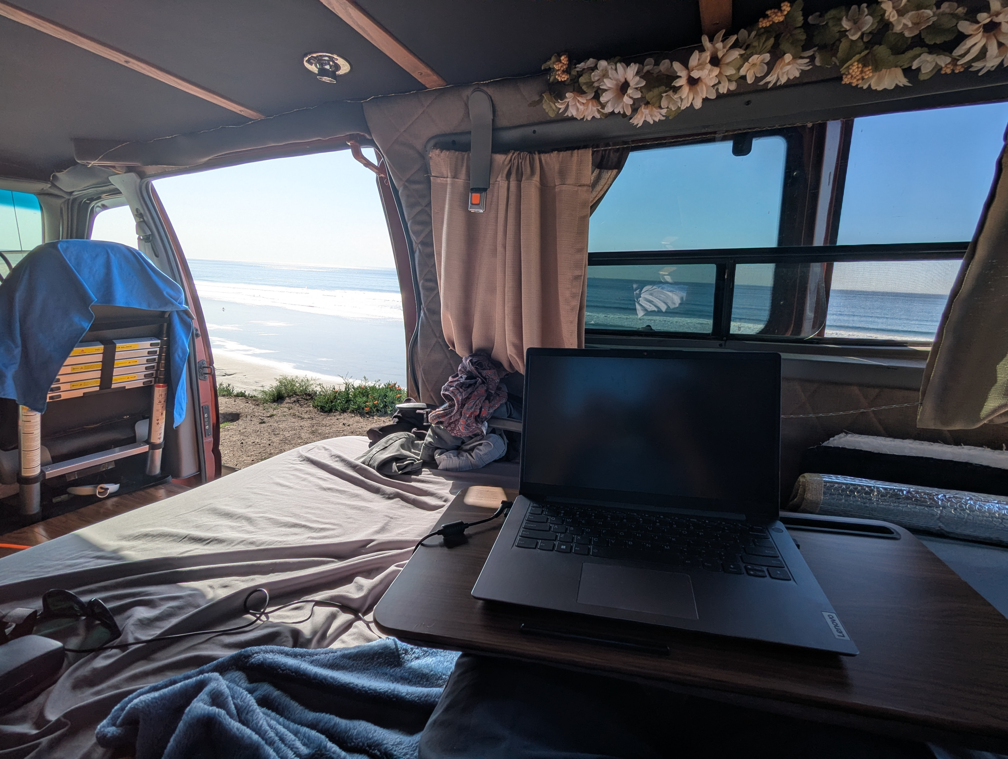 Inside an **RV rental** van, a laptop is set up for remote work on a bed. The open door and window reveal a sunny **beach camping** view.. Ford Econoline 1998