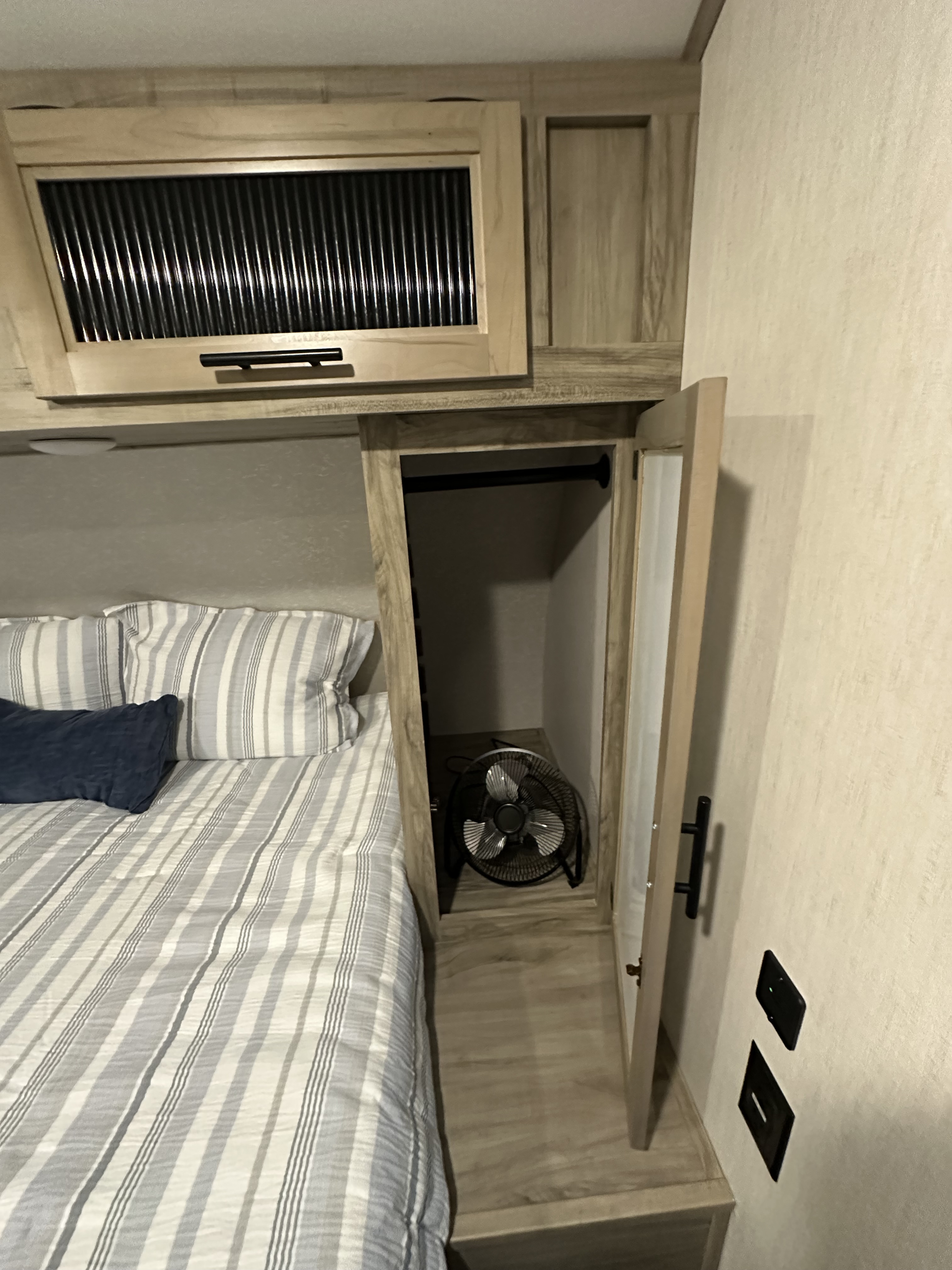Cozy RV rental bedroom with striped bedding, overhead cabinet, and an open closet revealing a fan for comfort.. Forest River Cherokee Alpha Wolf 2021