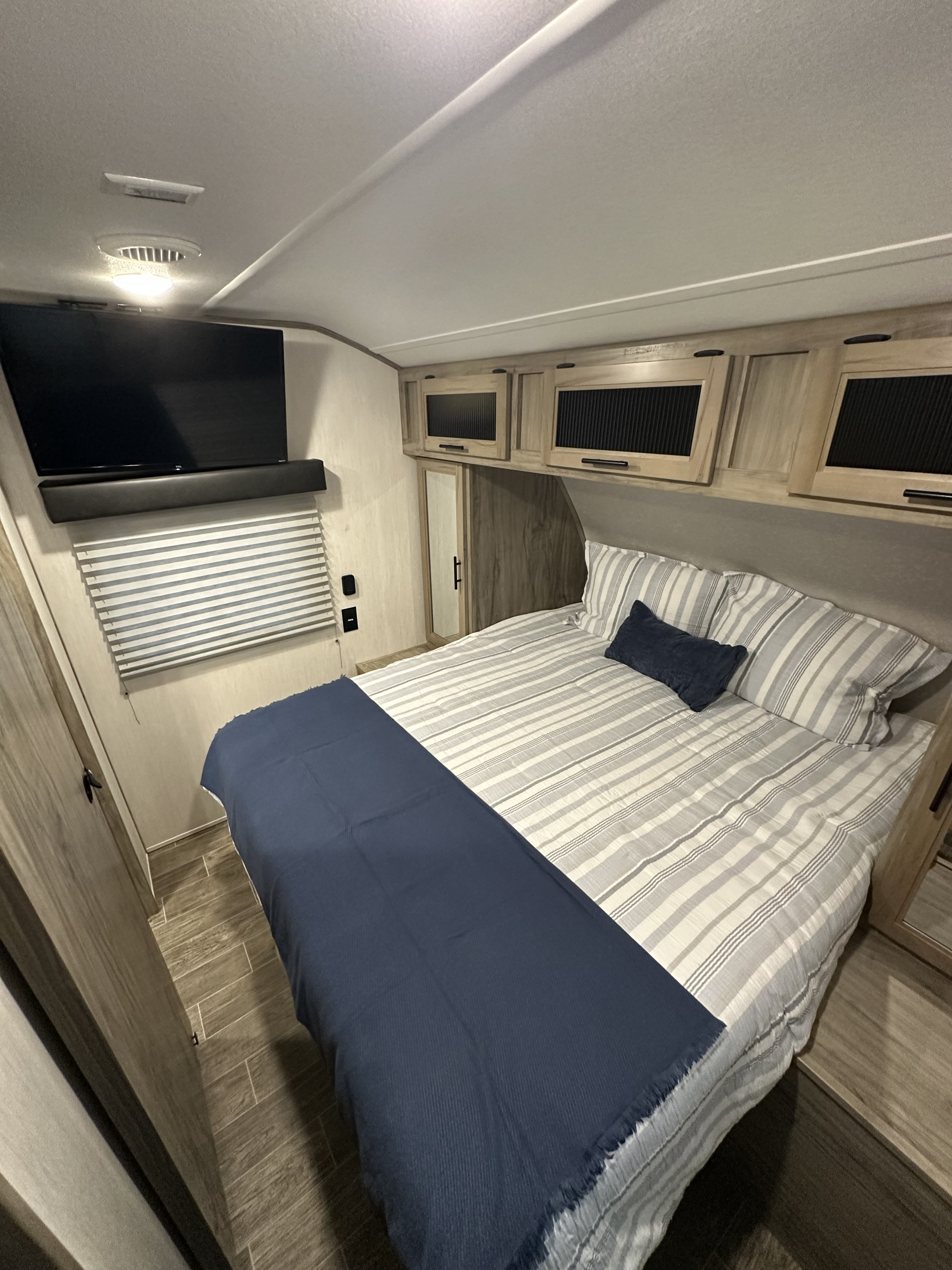 RV rental bedroom featuring a striped bed with a blue throw, wall-mounted TV, soundbar, and overhead storage cabinets.. Forest River Cherokee Alpha Wolf 2021