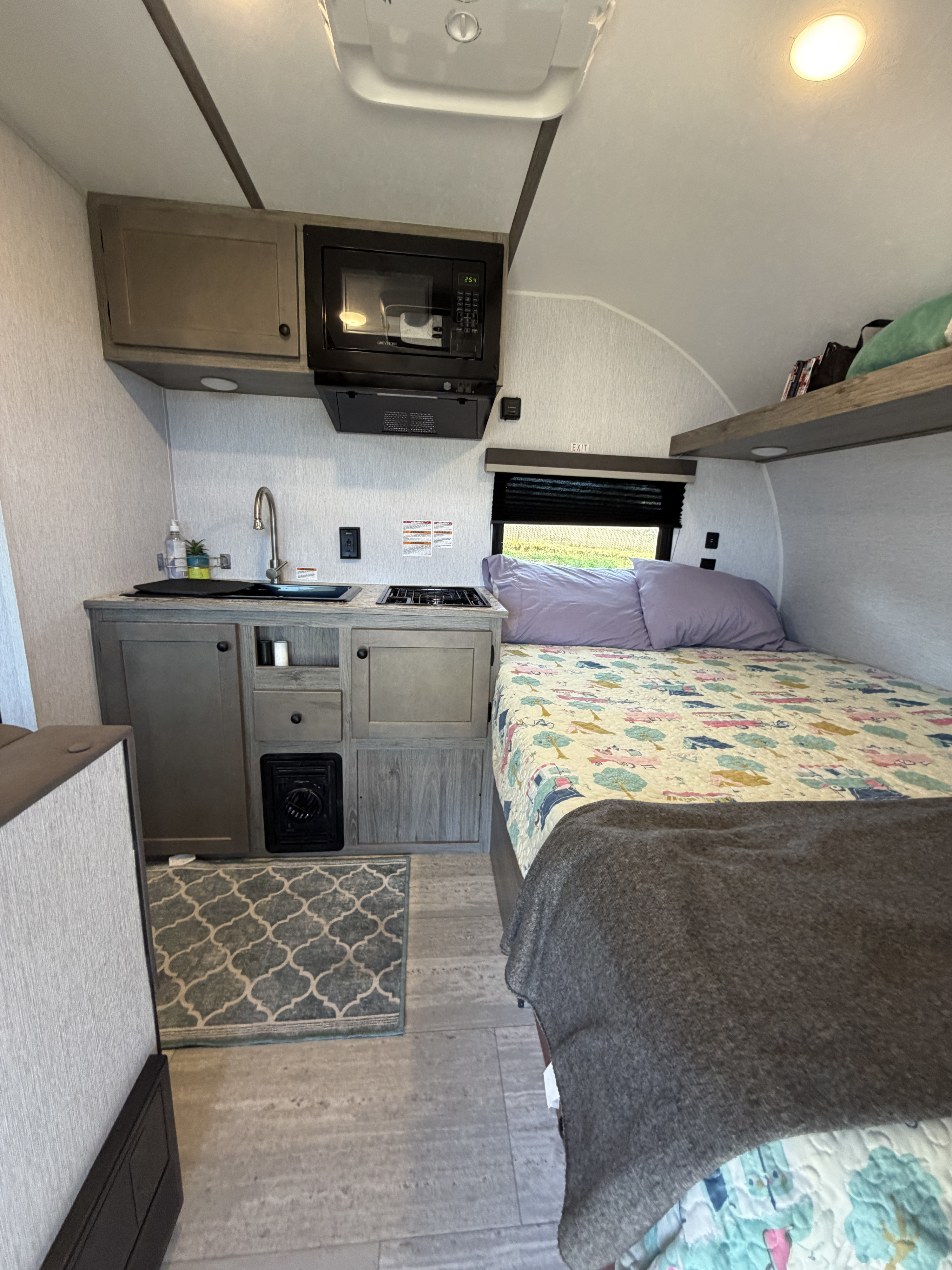 RV rental interior: compact kitchen with sink, microwave, stove, & cabinets, adjacent to a bed with a colorful quilt.. Sunset Park RV SunRay 2025
