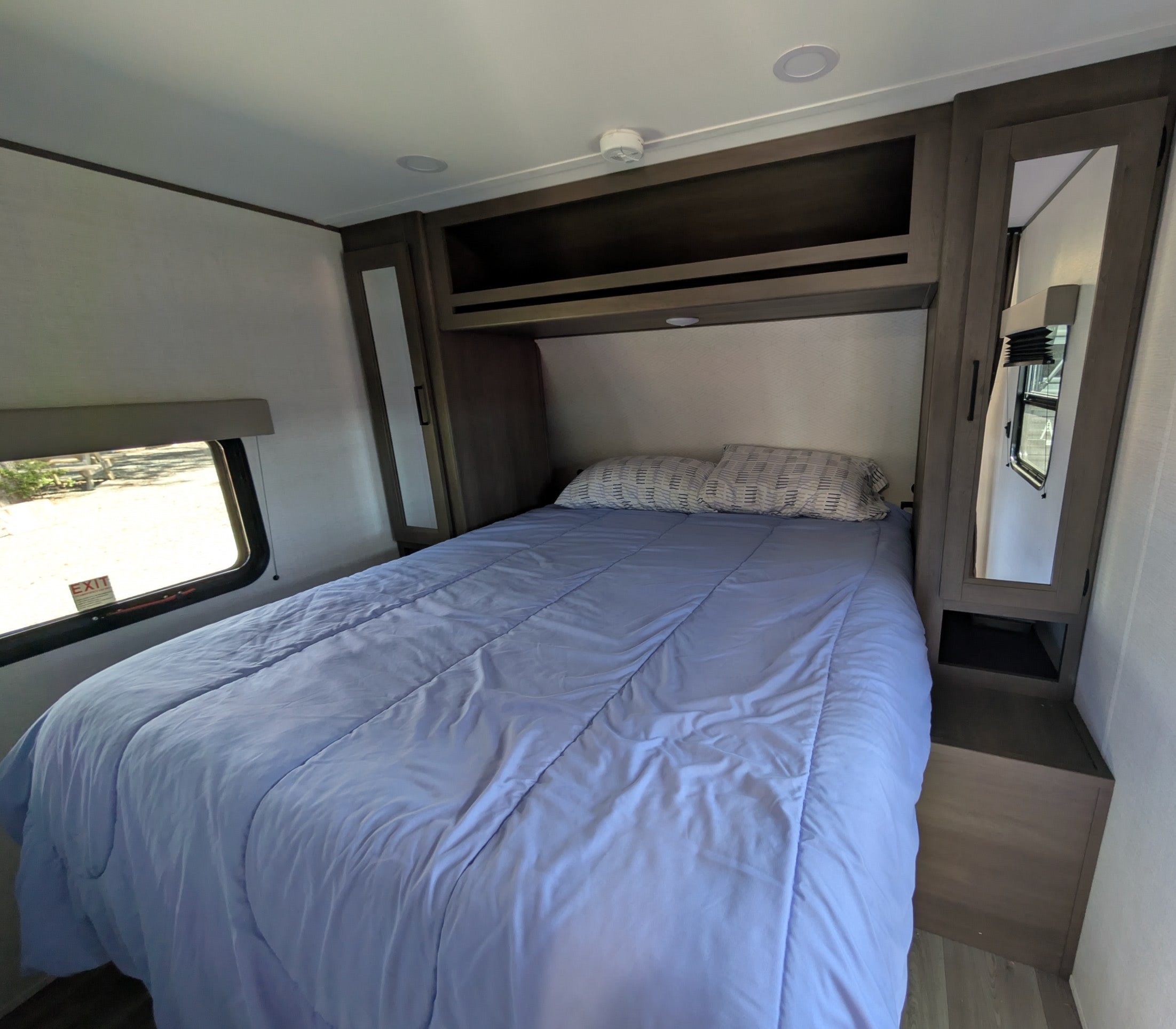 RV rental bedroom with a bed, overhead storage, mirrored side cabinets, and a side escape window.. Grand Design Transcend Xplor 2025