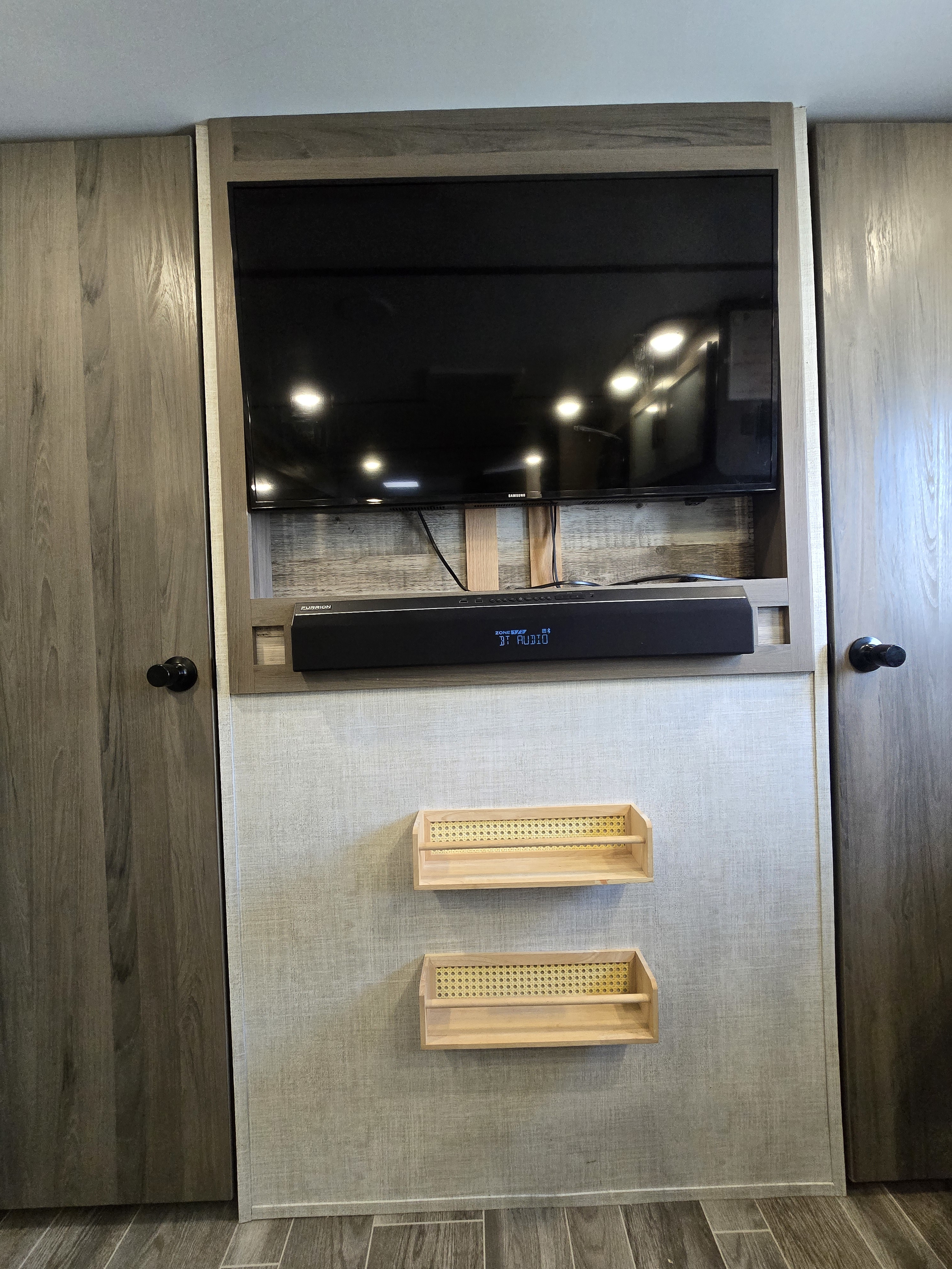 RV rental interior featuring a wall-mounted TV, soundbar, and two small shelves, flanked by wood-grain cabinet doors.. Gulf Stream Kingsport 2022