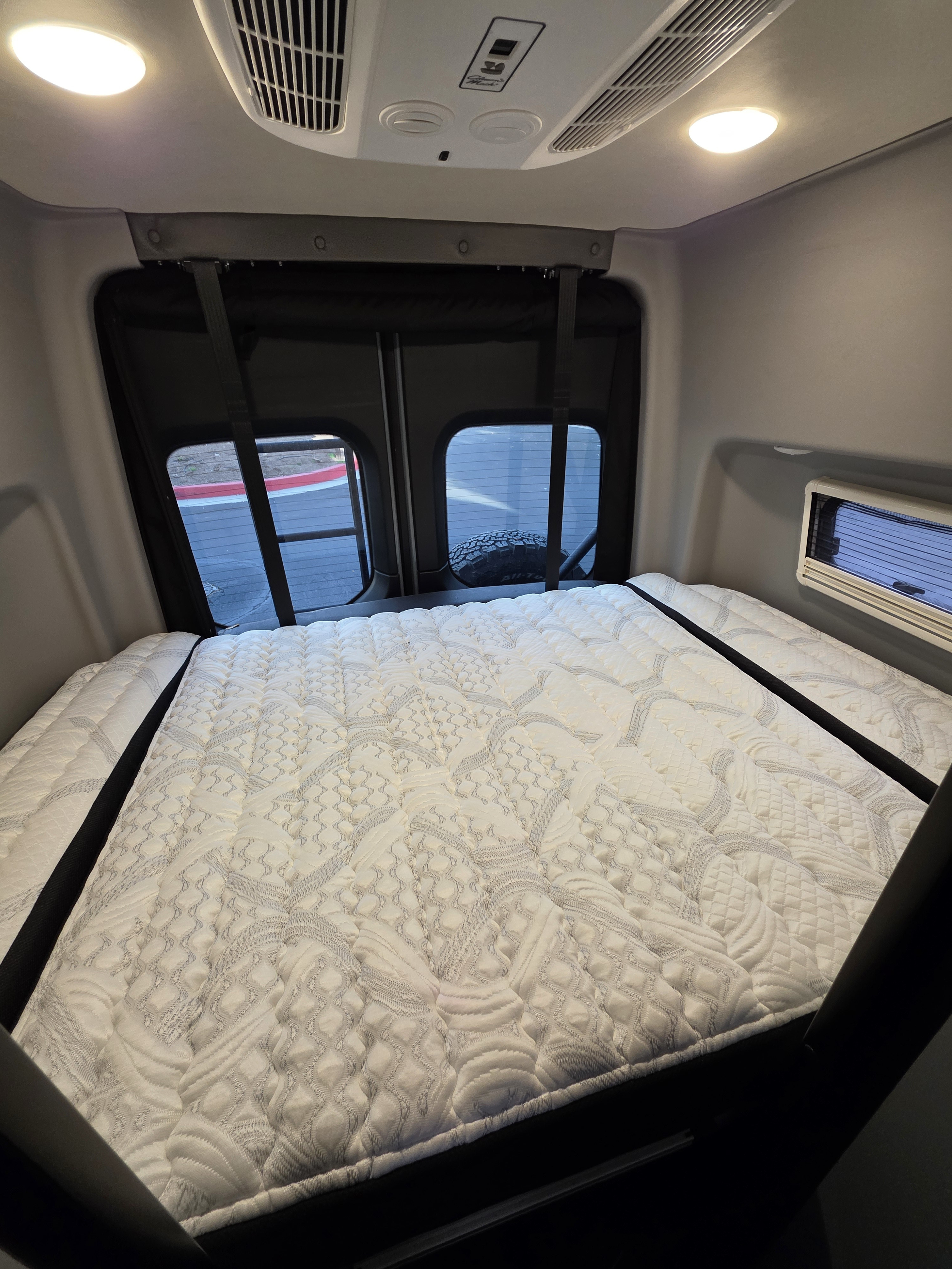 Inside an RV rental: a plush, quilted mattress fills the rear sleeping area, flanked by windows. Ceiling features lights & AC.. Winnebago Revel 2022