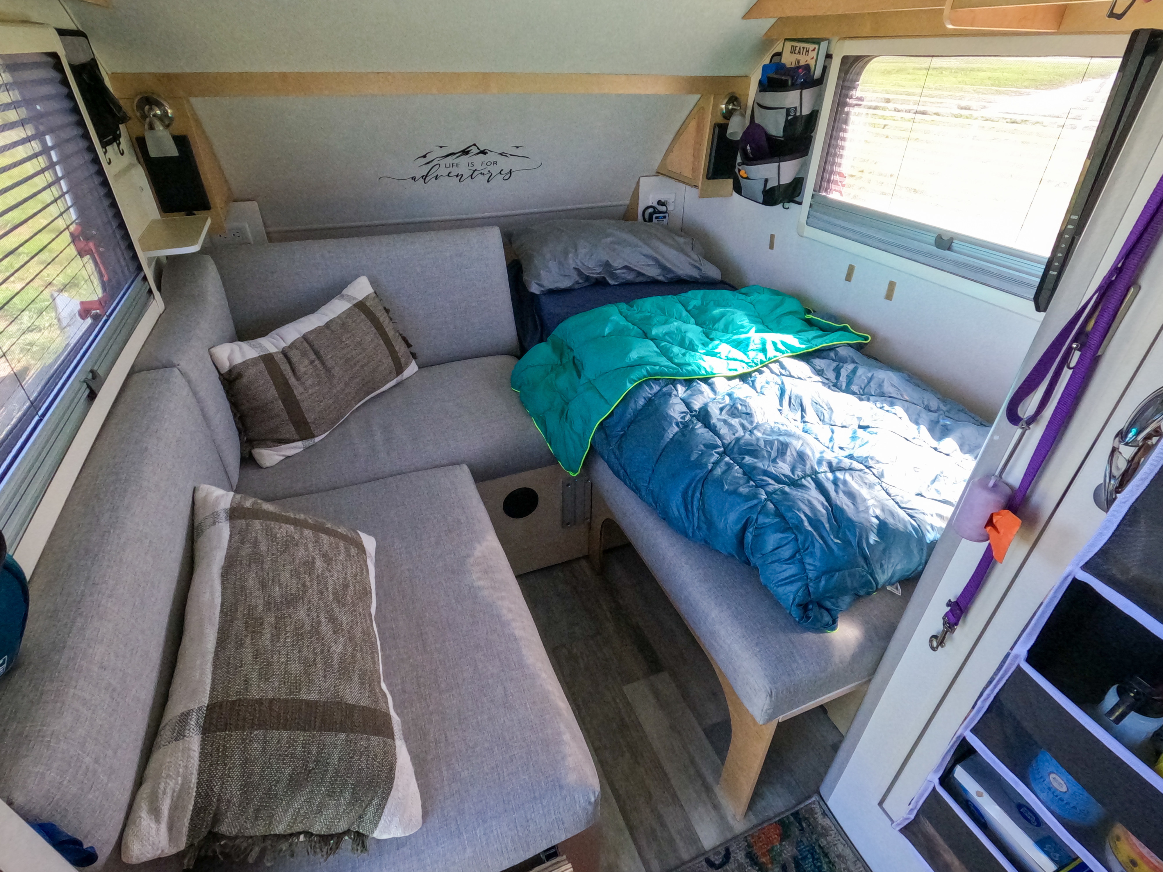 This RV rental's interior features comfy gray seating, a bed with blue bedding, and "Life is for adventures" wall art.. nuCamp T@B 2022