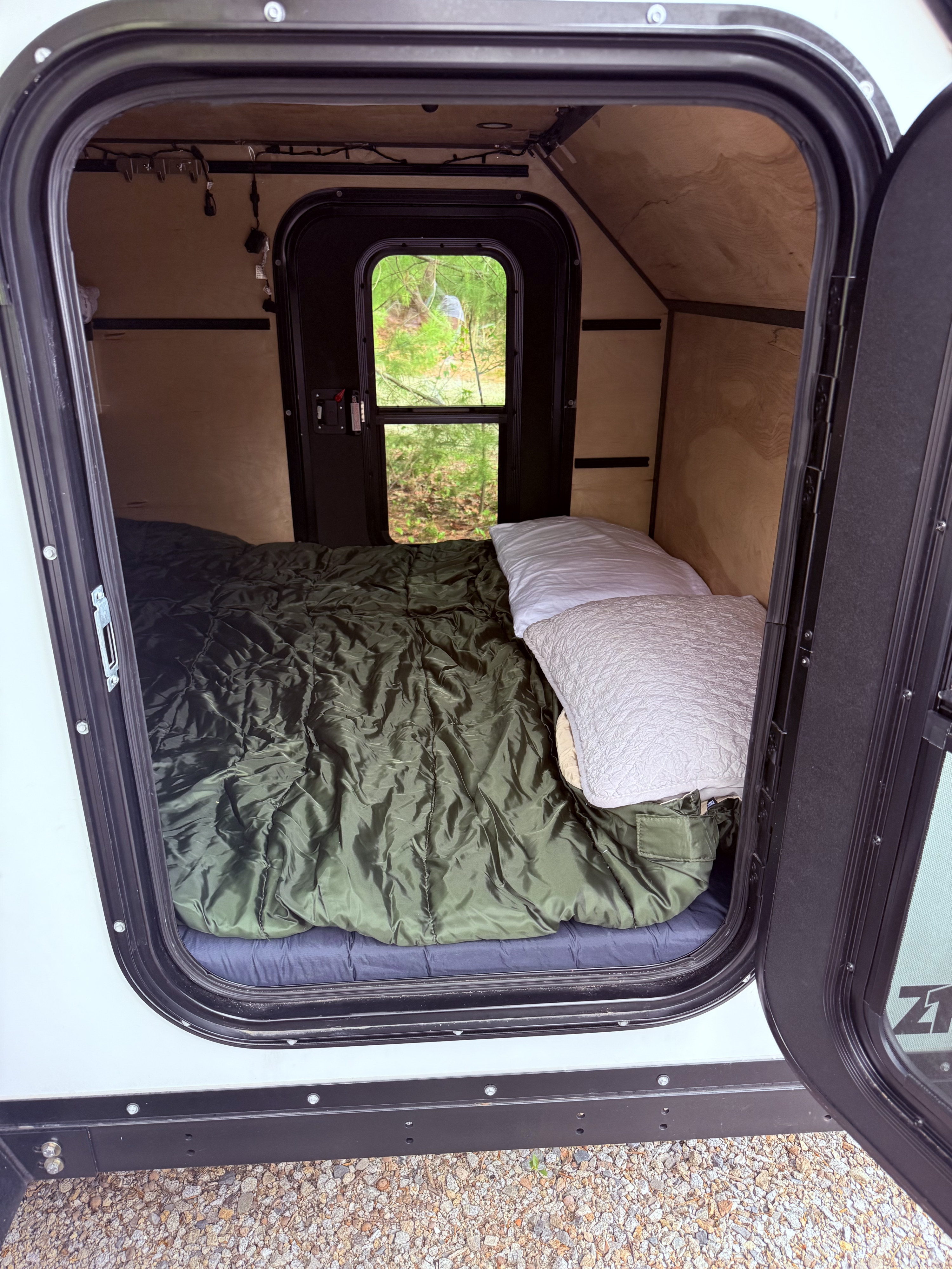 A cozy RV rental interior features a bed with green bedding and white pillows. A window reveals lush green trees outside.. Zion Off Road Z1 2025