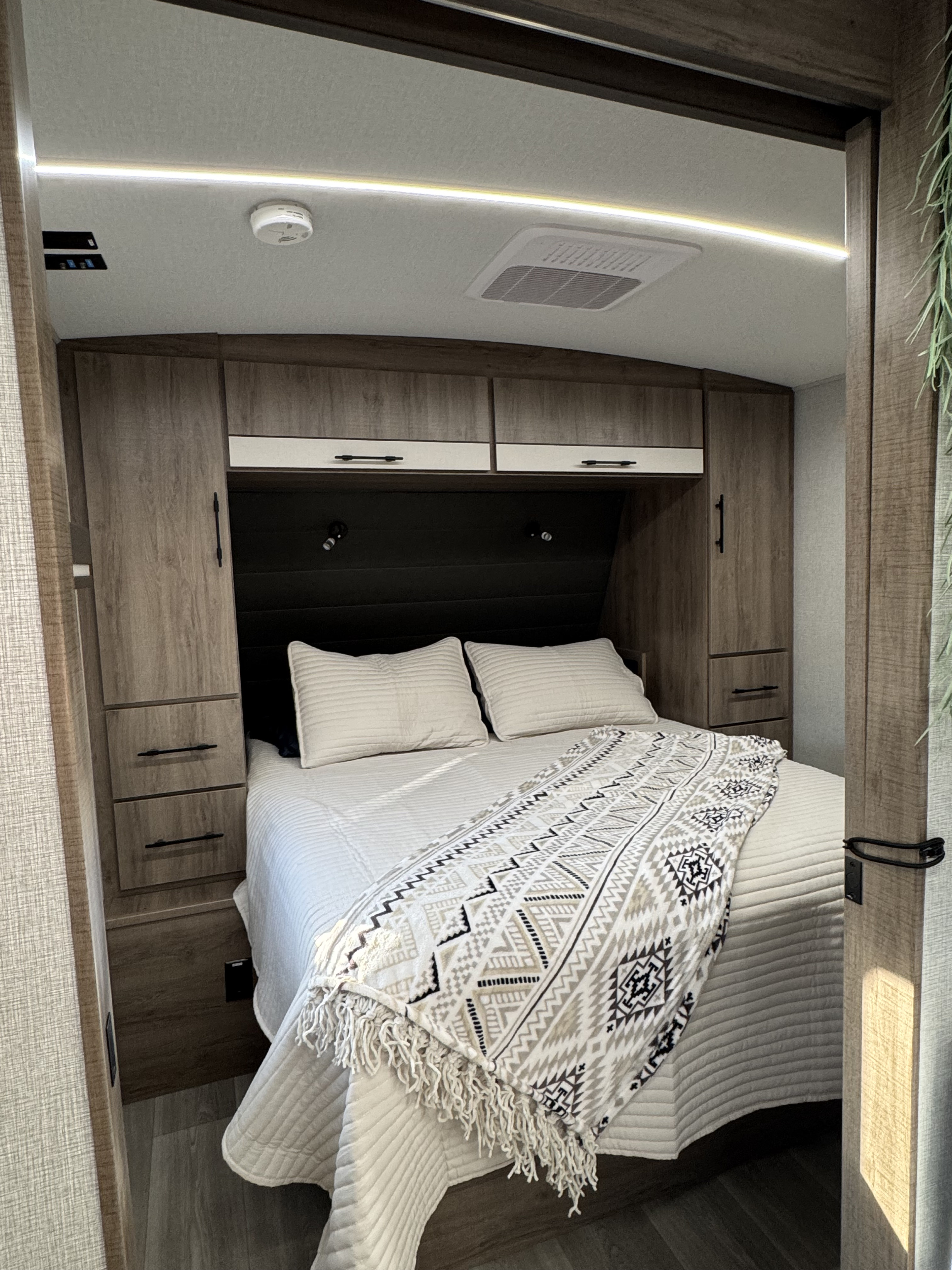 Relax in this inviting RV rental bedroom, featuring a comfortable bed with a patterned throw, ample wood storage, AC, and accent lighting.. Grand Design Imagine 2025