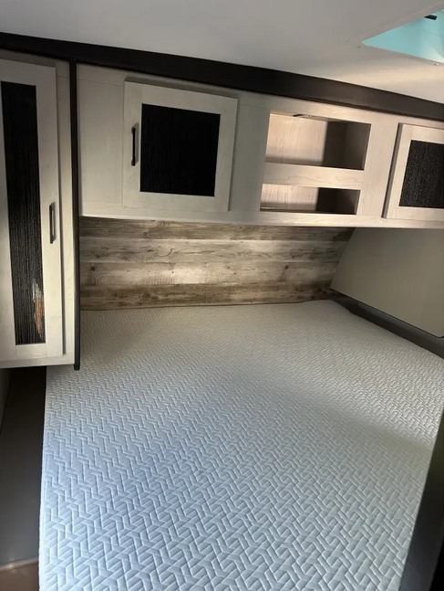 Inside an RV rental: a cozy bedroom with a patterned queen mattress, overhead storage, and rustic wood paneling.. Heartland RVs Mallard 2022