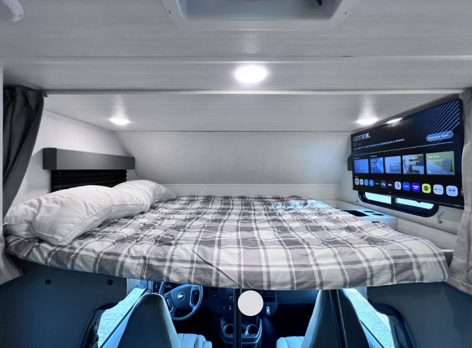This RV rental features a cozy overhead bed with plaid bedding, pillows, and a wall-mounted TV. The driver's cabin is visible below.. East to West Entrada 2026