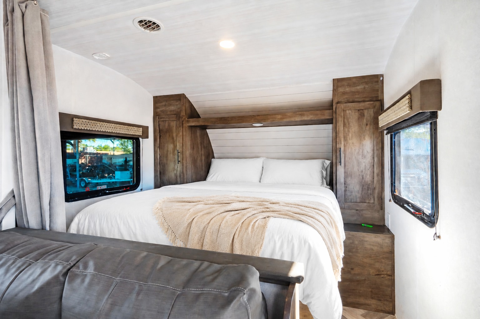Interior of an RV rental bedroom with a large, neatly made bed, wooden cabinets, and windows on both sides. A grey couch is in the foreground.. Forest River Wildwood X-Lite 2020