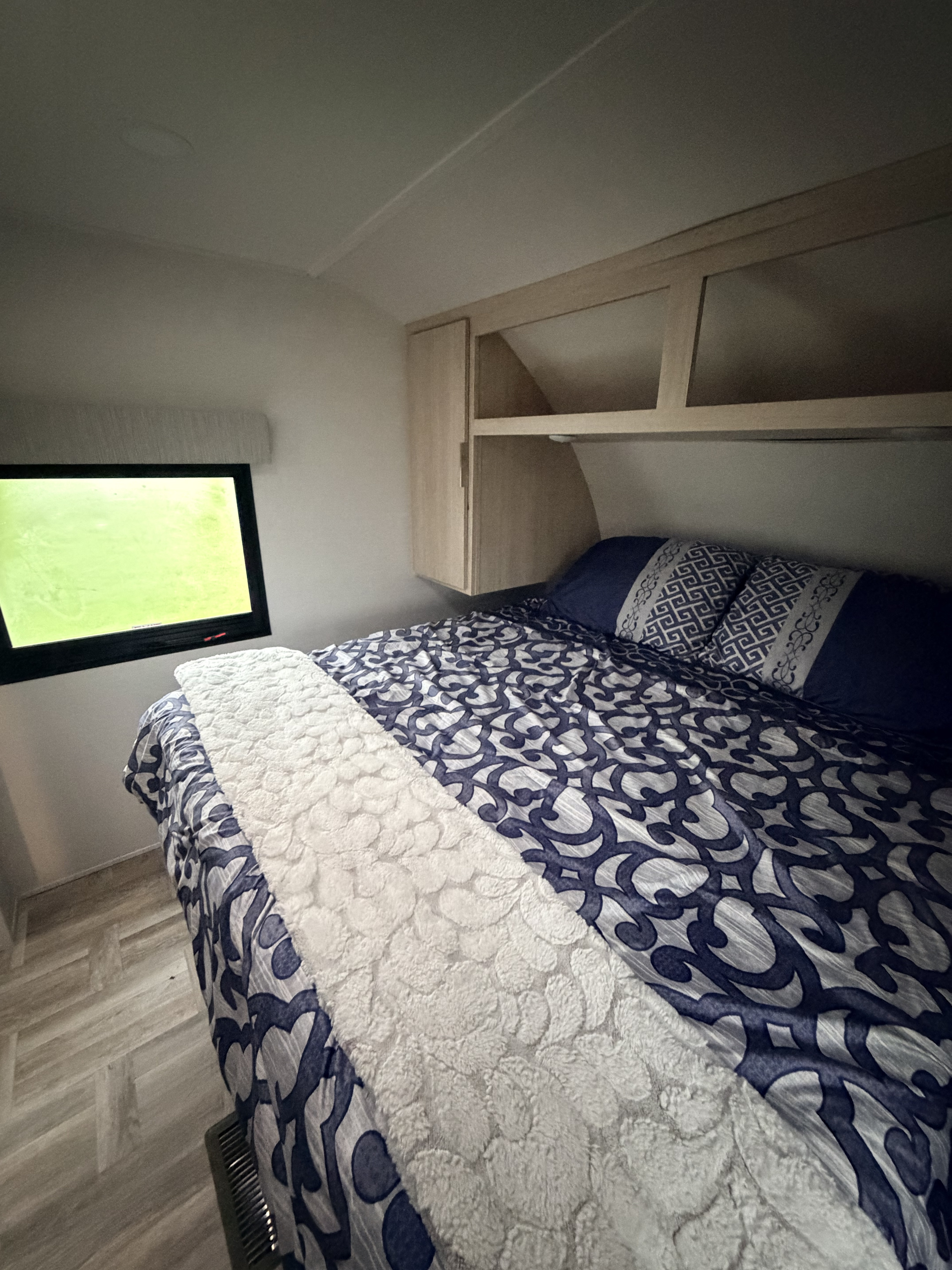 Cozy RV rental bedroom featuring blue patterned bedding, a white throw, overhead cabinets, and a window with green light.. Forest River Tracer Ultra Lite 2025