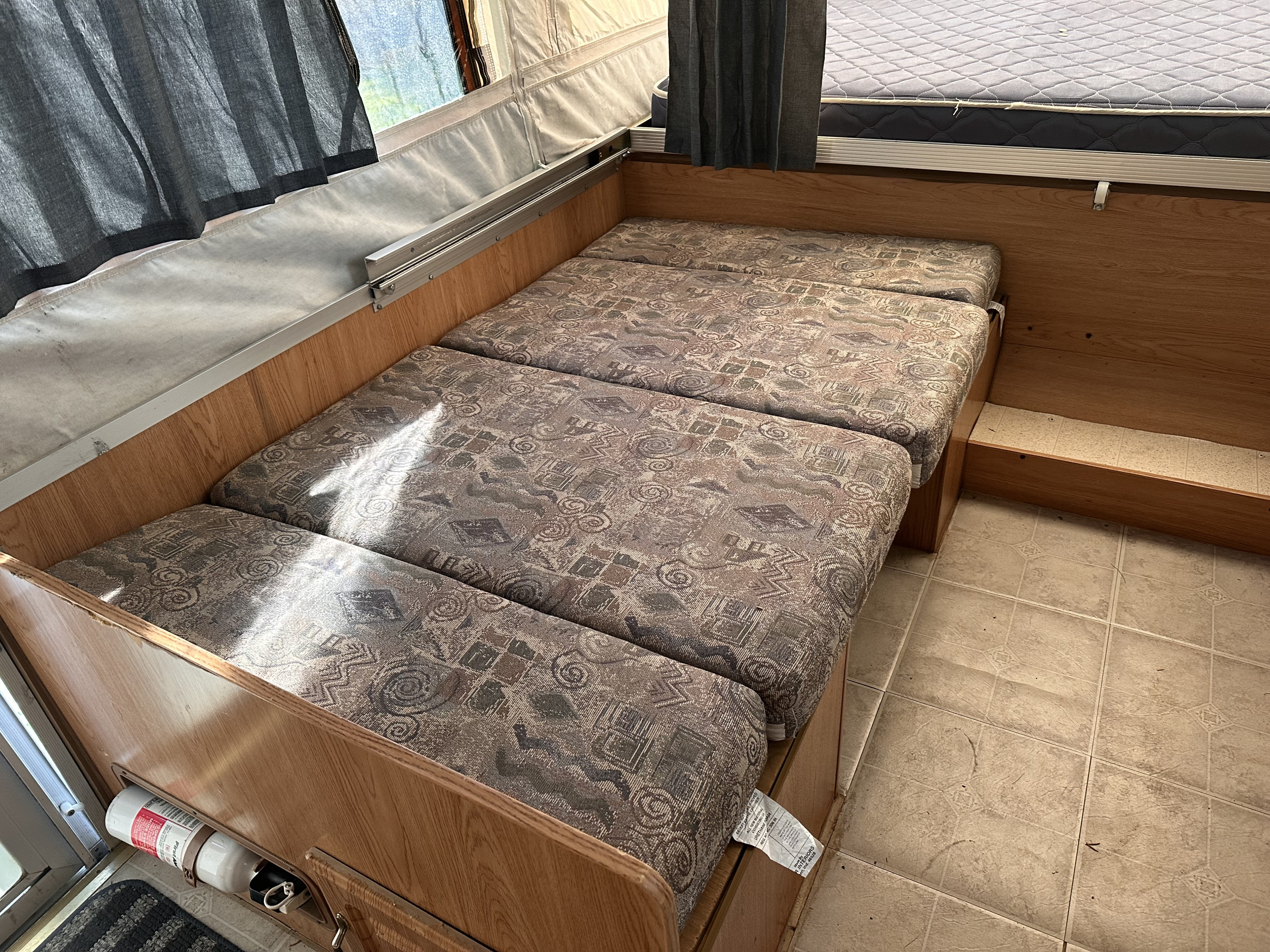 RV rental interior: A dinette with patterned cushions, convertible to a bed, with gray curtains and a fire extinguisher.. Forest River Rockwood Premier 2004