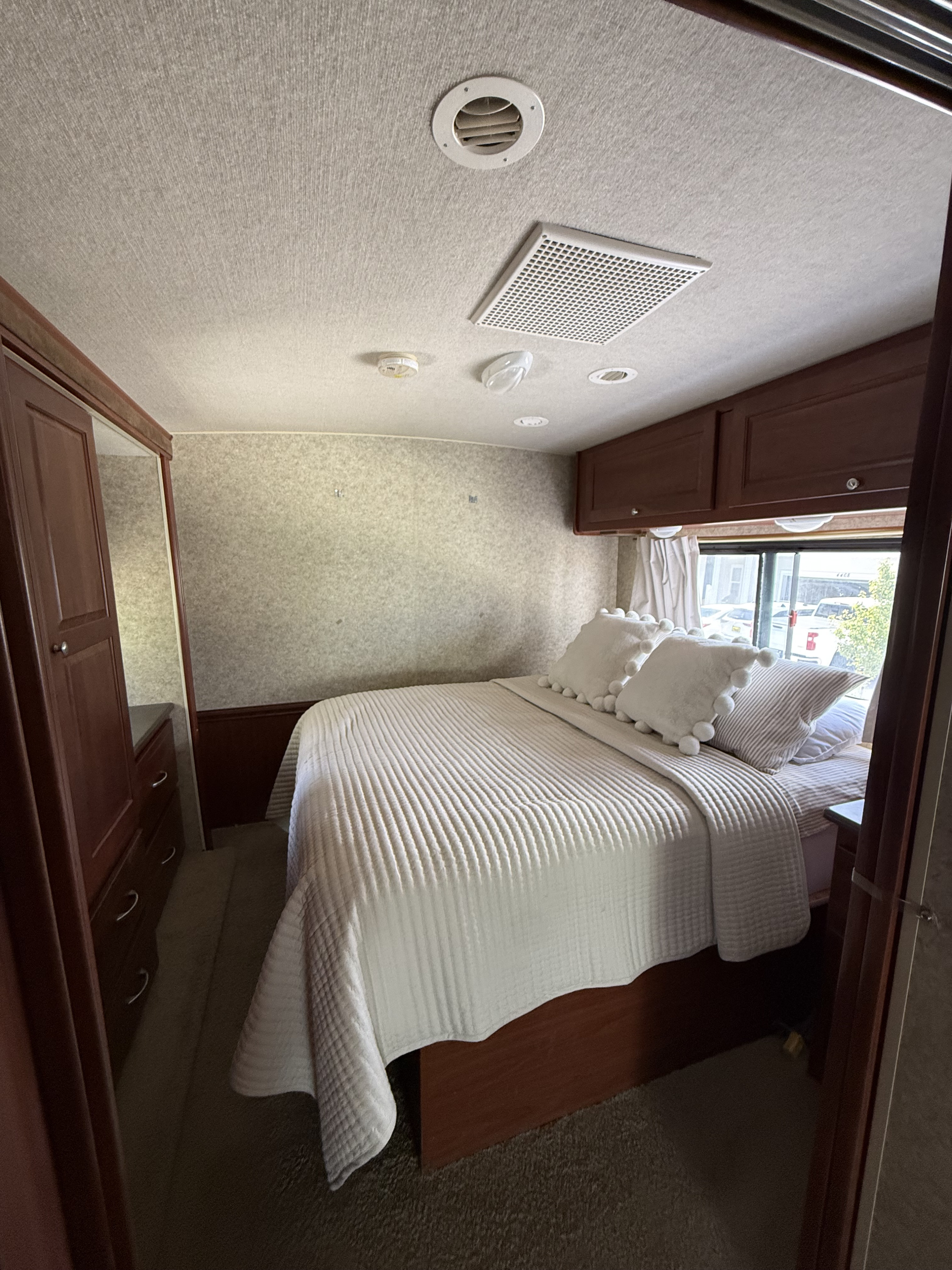 Cozy RV rental bedroom with comfy queen bed, white bedding, dark wood storage, and bright window view.. Itasca Sunstar 2011