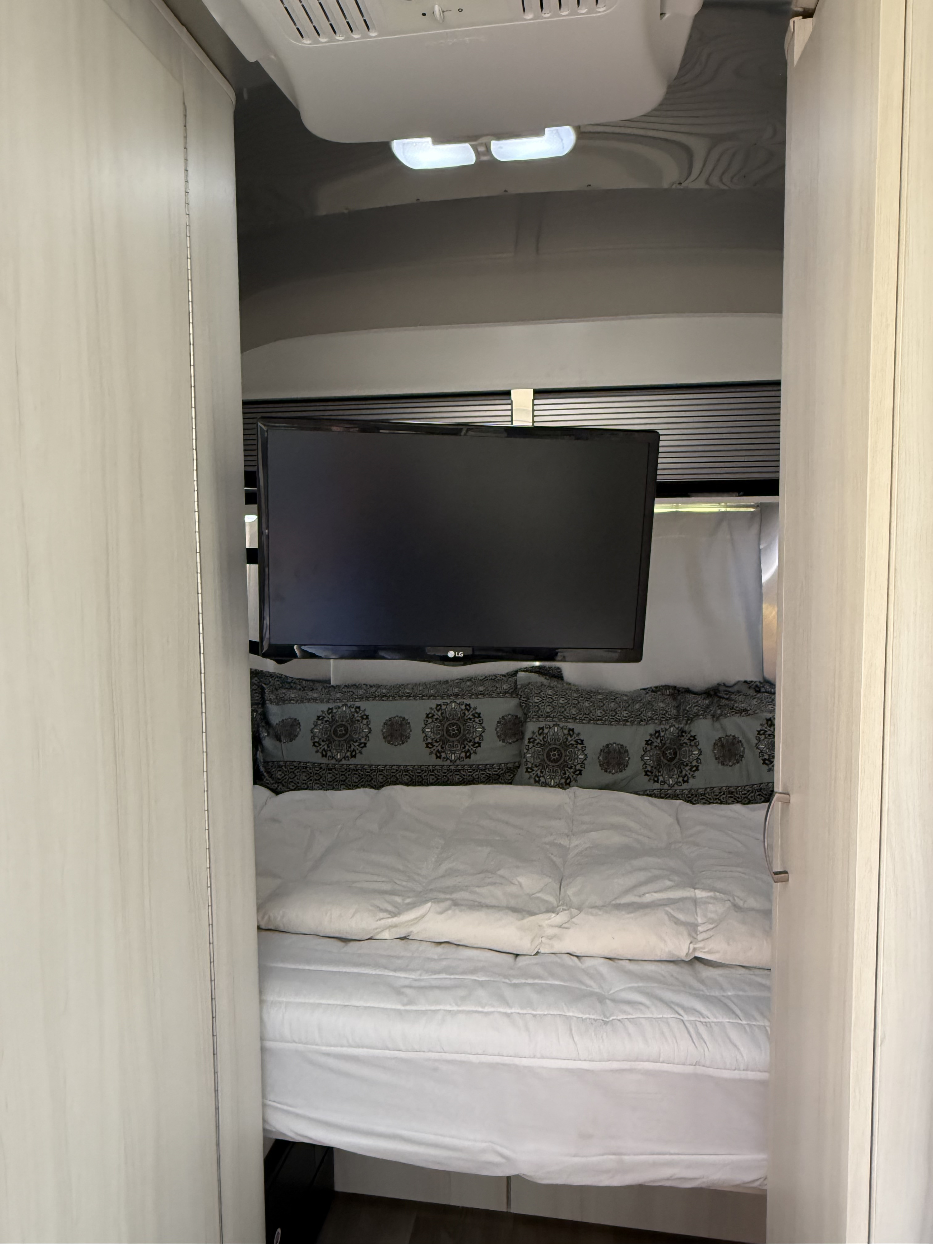 RV rental interior: a compact bedroom features a mounted LG TV, a cozy bed with decorative pillows, and an overhead AC unit.. Airstream Sport 2019