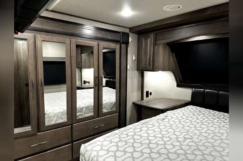 RV rental bedroom with modern dark wood finishes, comfy bed, mirrored wardrobe, and TV, offering a cozy retreat on the road.. Grand Design Solitude S-Class 2022