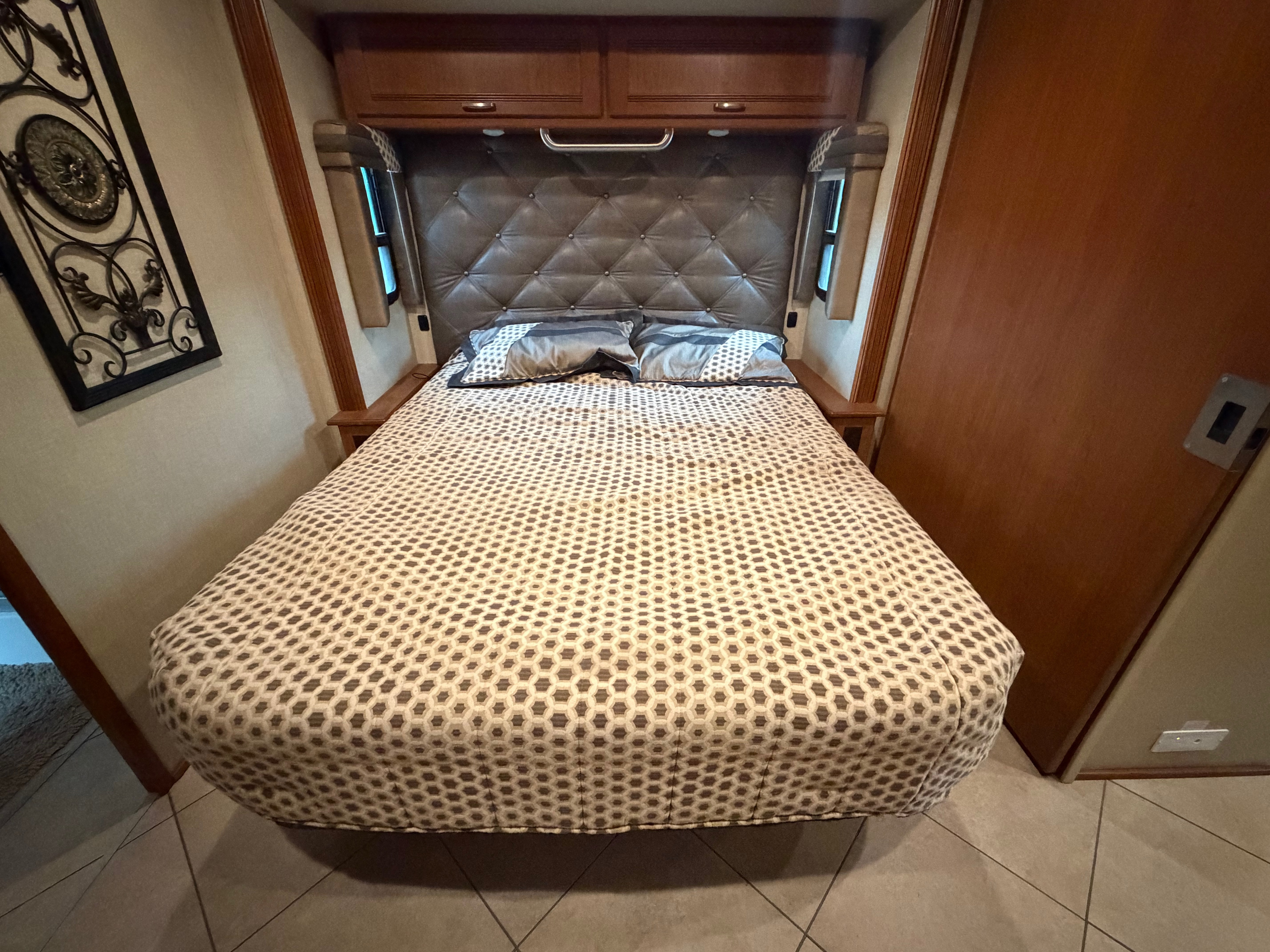 Cozy RV rental bedroom featuring a geometric bedspread, tufted headboard, overhead storage, and small windows. Ideal for travel accommodations.. Winnebago Forza 2014