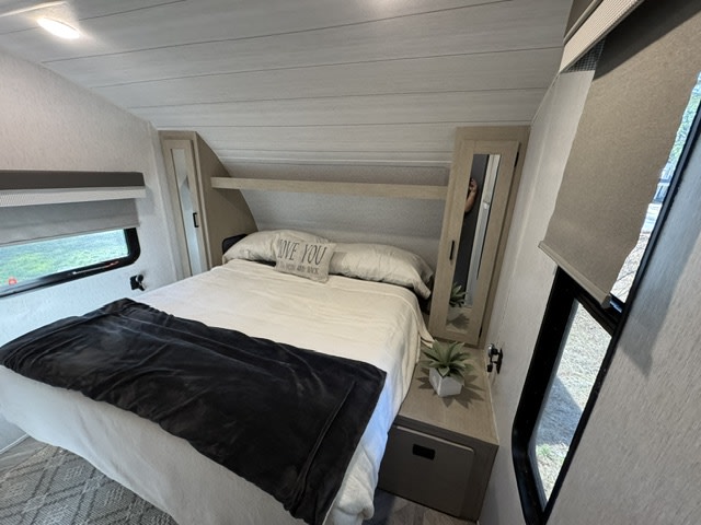 This cozy RV rental bedroom features a comfortable bed with a throw, mirrored cabinet, windows, and overhead storage for travel comfort.. Forest River Wildwood X-Lite 2023