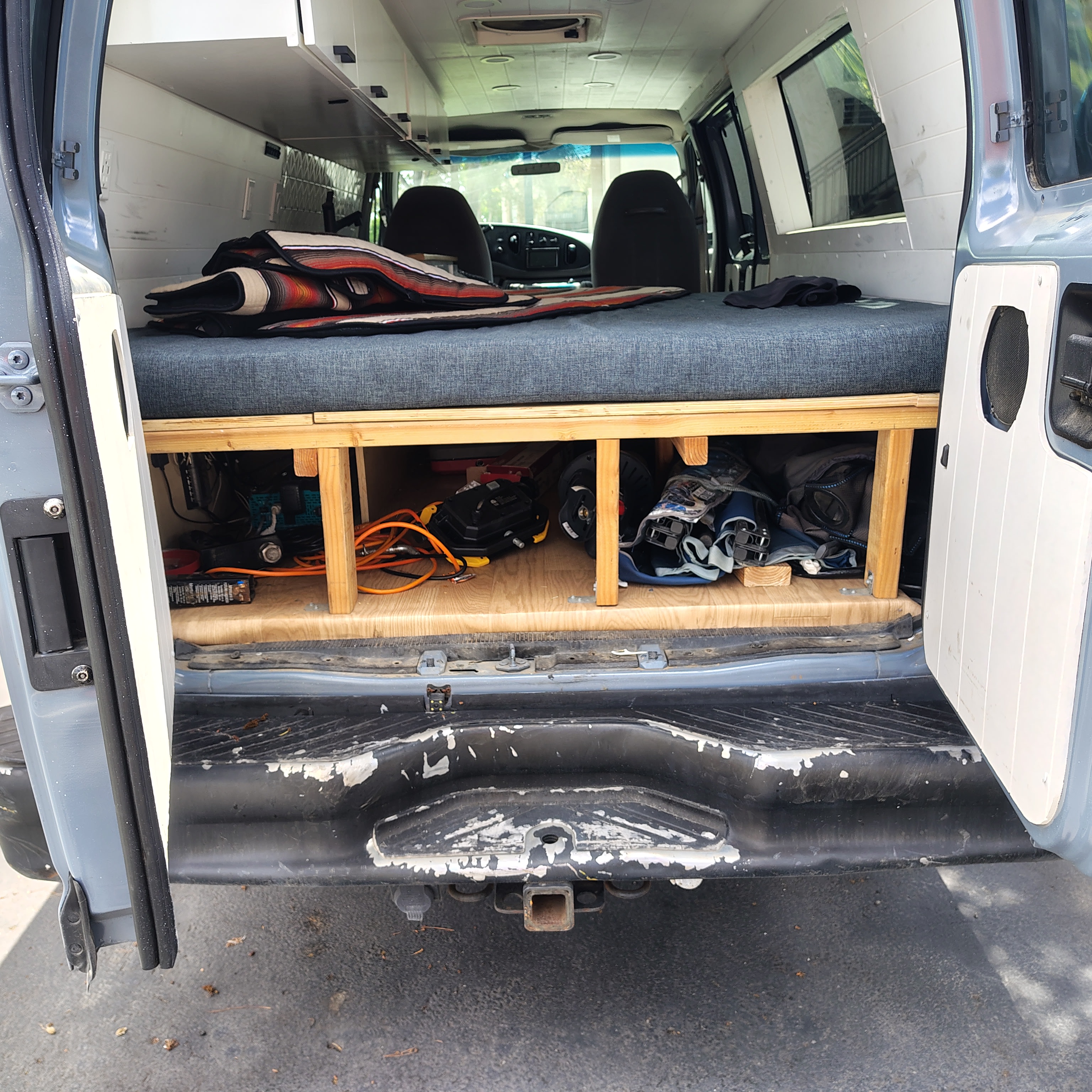 RV rental camper van interior: A blue bed cushion on a wooden platform with tools and bags stored below, seen through open rear doors.. Ford Econoline E350 2001