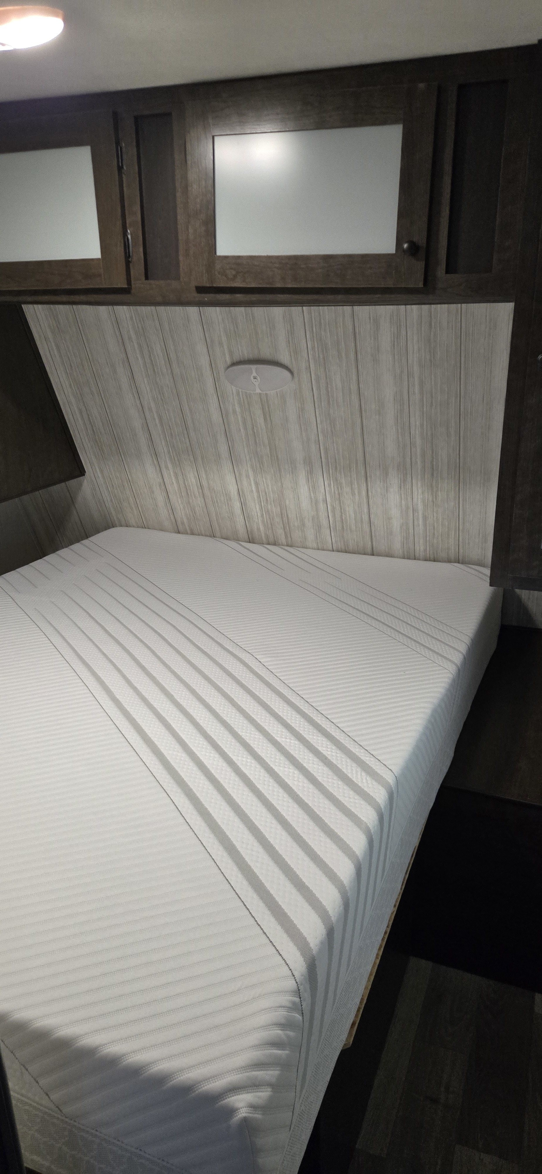 Inside an RV rental: a cozy bedroom features a striped white mattress, wood-paneled headboard, and overhead cabinets.. Heartland RVs Pioneer 2020