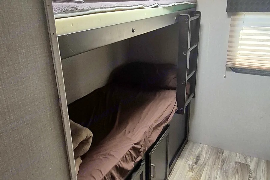 This RV rental features cozy bunk beds; the lower bunk has brown bedding, storage below, and a window with blinds.. Venture Sporttrek 2019