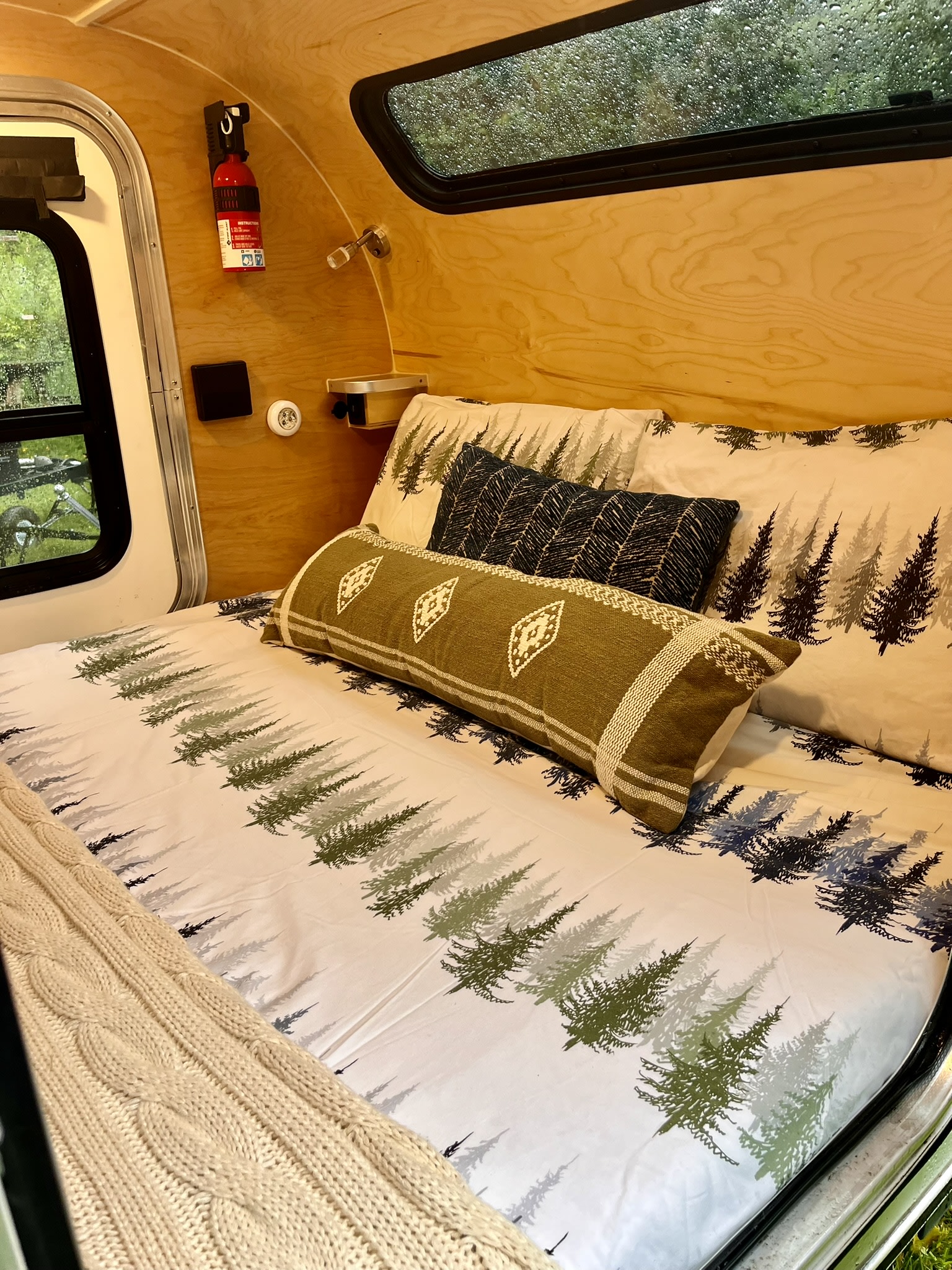 Cozy RV rental interior features a bed with white forest-print bedding, decorative pillows, and a knit blanket. Wood-paneled walls.. Aero Steel 2022