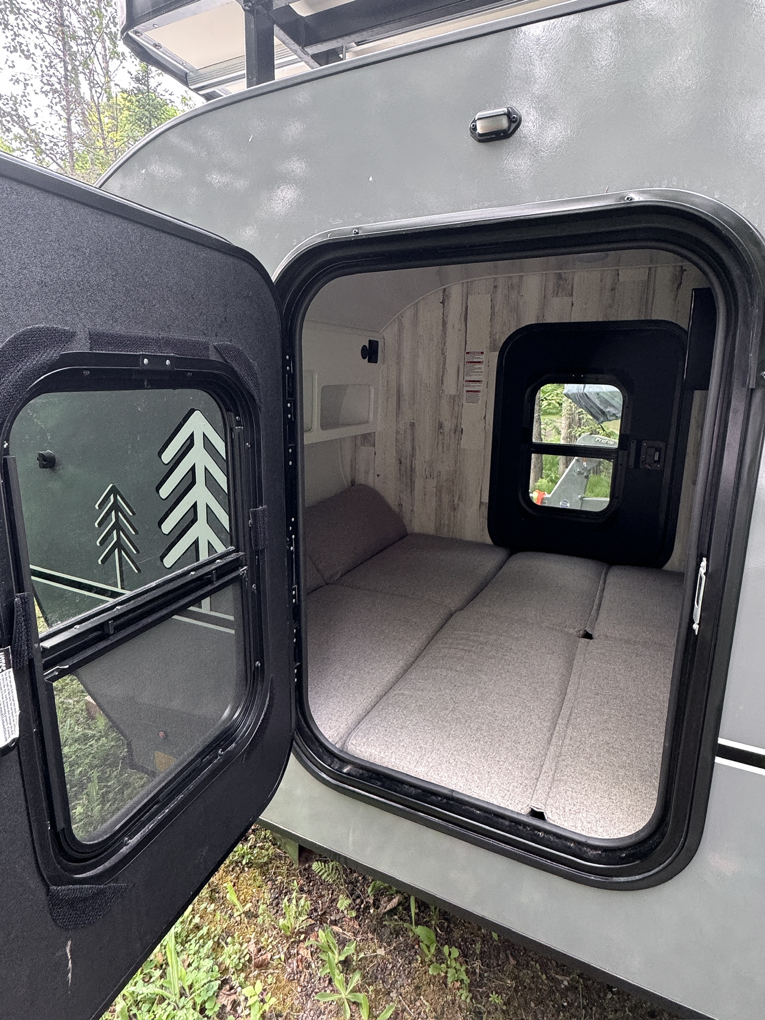 Open door of a gray rv rental, revealing a cozy interior with a gray cushion bed, light wood walls, and a small window.. Modern Buggy RV CB12 2025