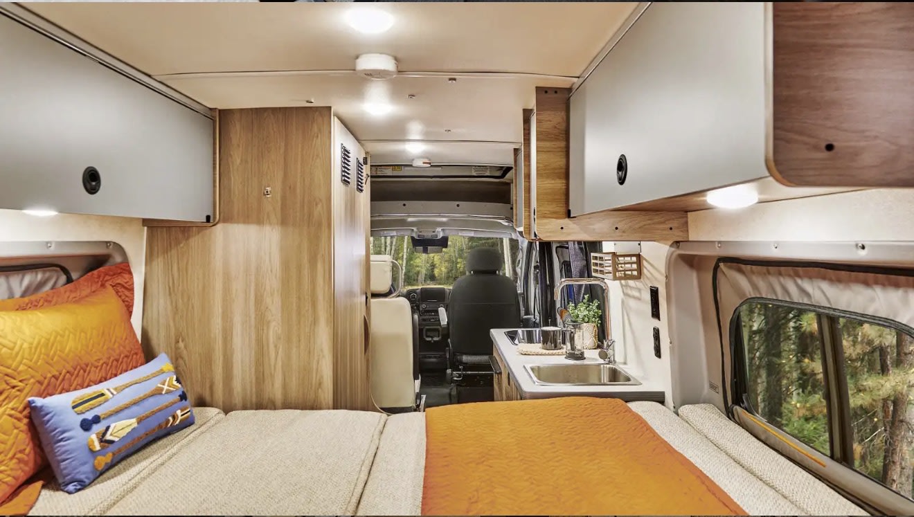 RV rental interior: Cozy camper van with bed, kitchenette, and overhead storage, looking forward to the driver's cabin.. Winnebago Solis 59P National Park Fundation 2026