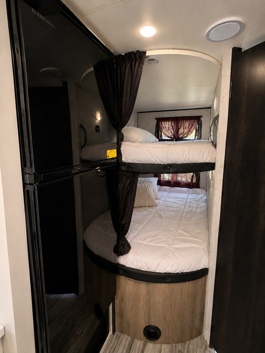 An RV rental's interior shows two bunk beds with white bedding & dark curtains. A tall refrigerator reflects the scene.. Jayco Jay Flight 2024