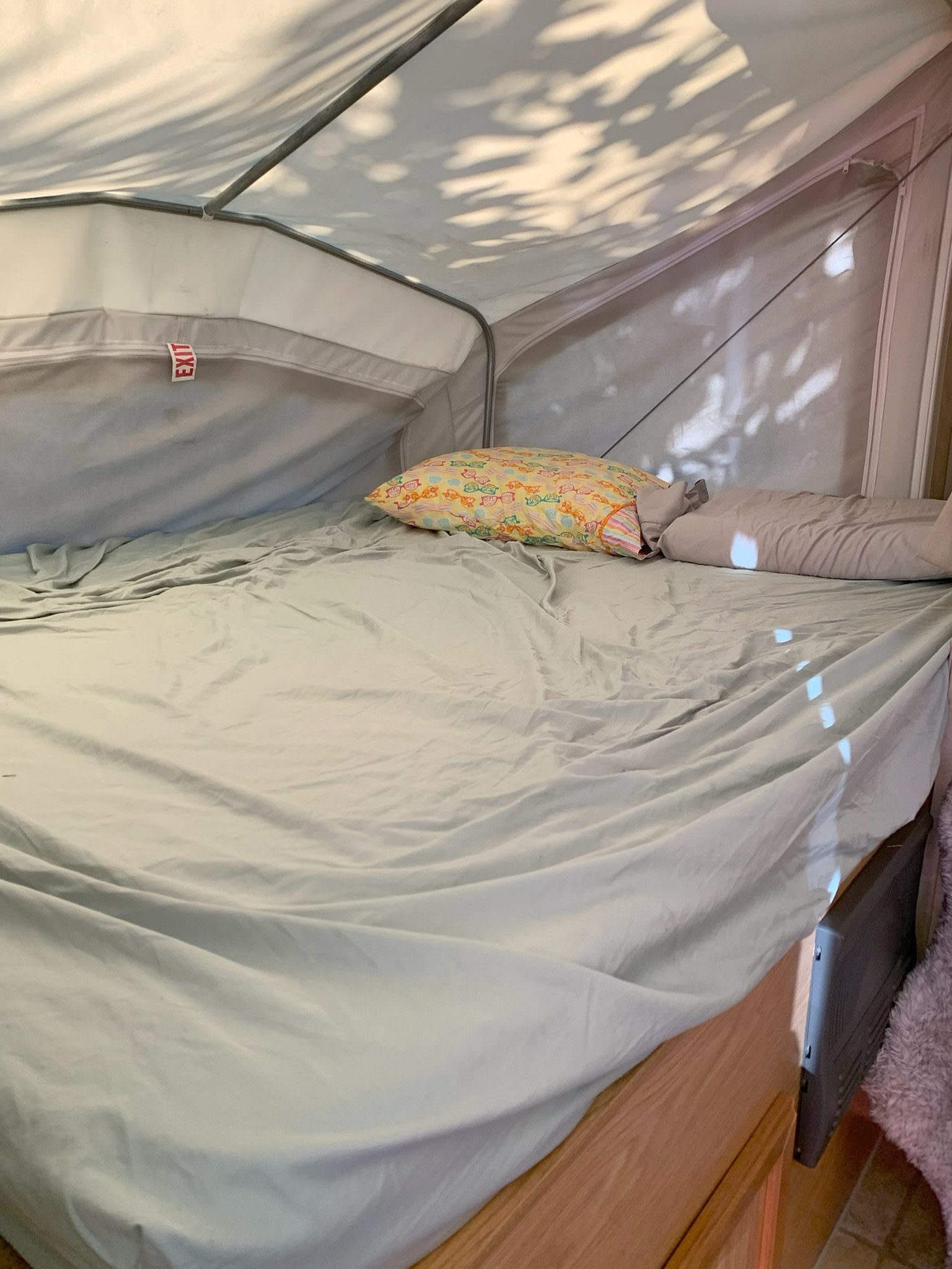 RV rental pop-up camper bed with light sheets, colorful pillows, and sun shadows on canvas walls. Exit tag visible.. KZ CCH 2005
