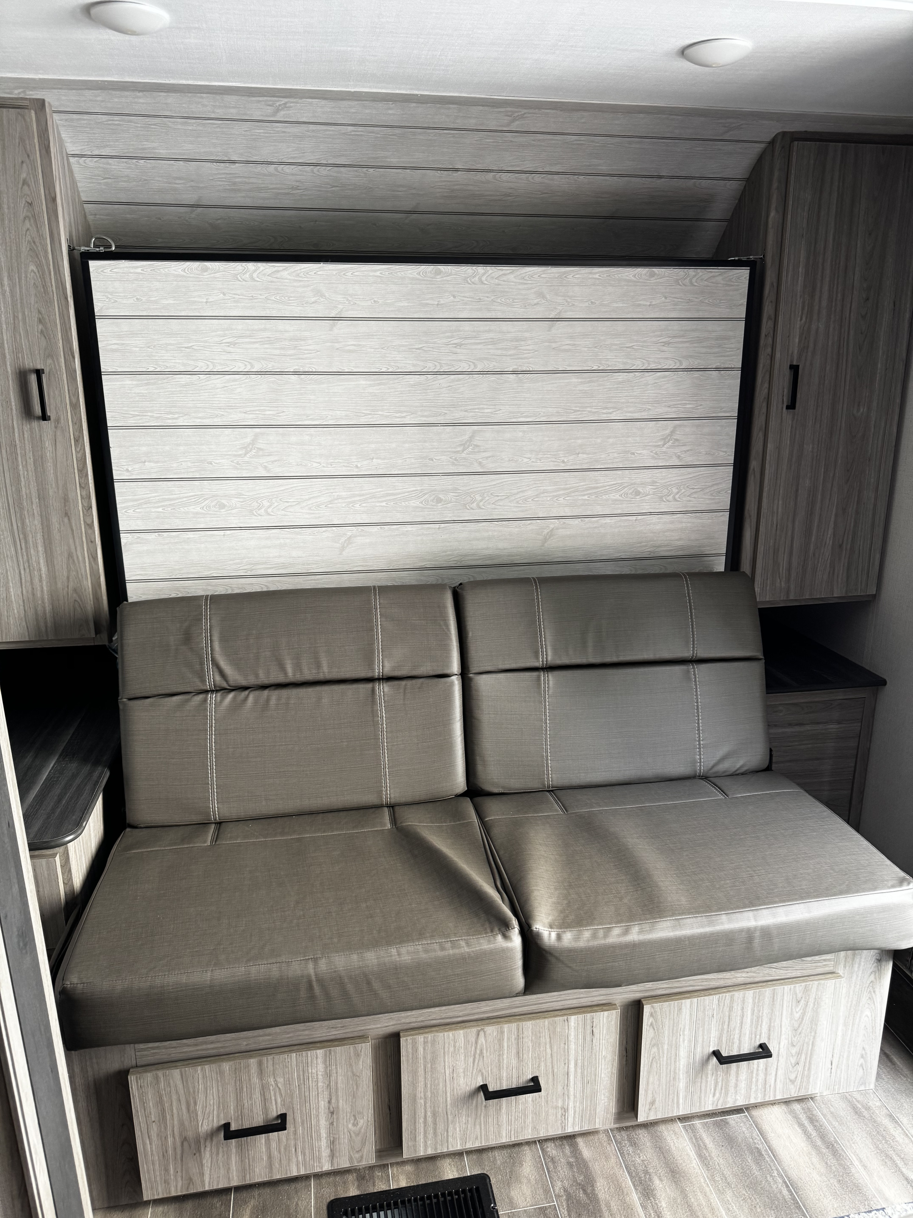 Modern rv rental interior featuring a gray sofa, light wood paneling, overhead cabinets, and under-sofa storage drawers.. Keystone RV Passport 2022