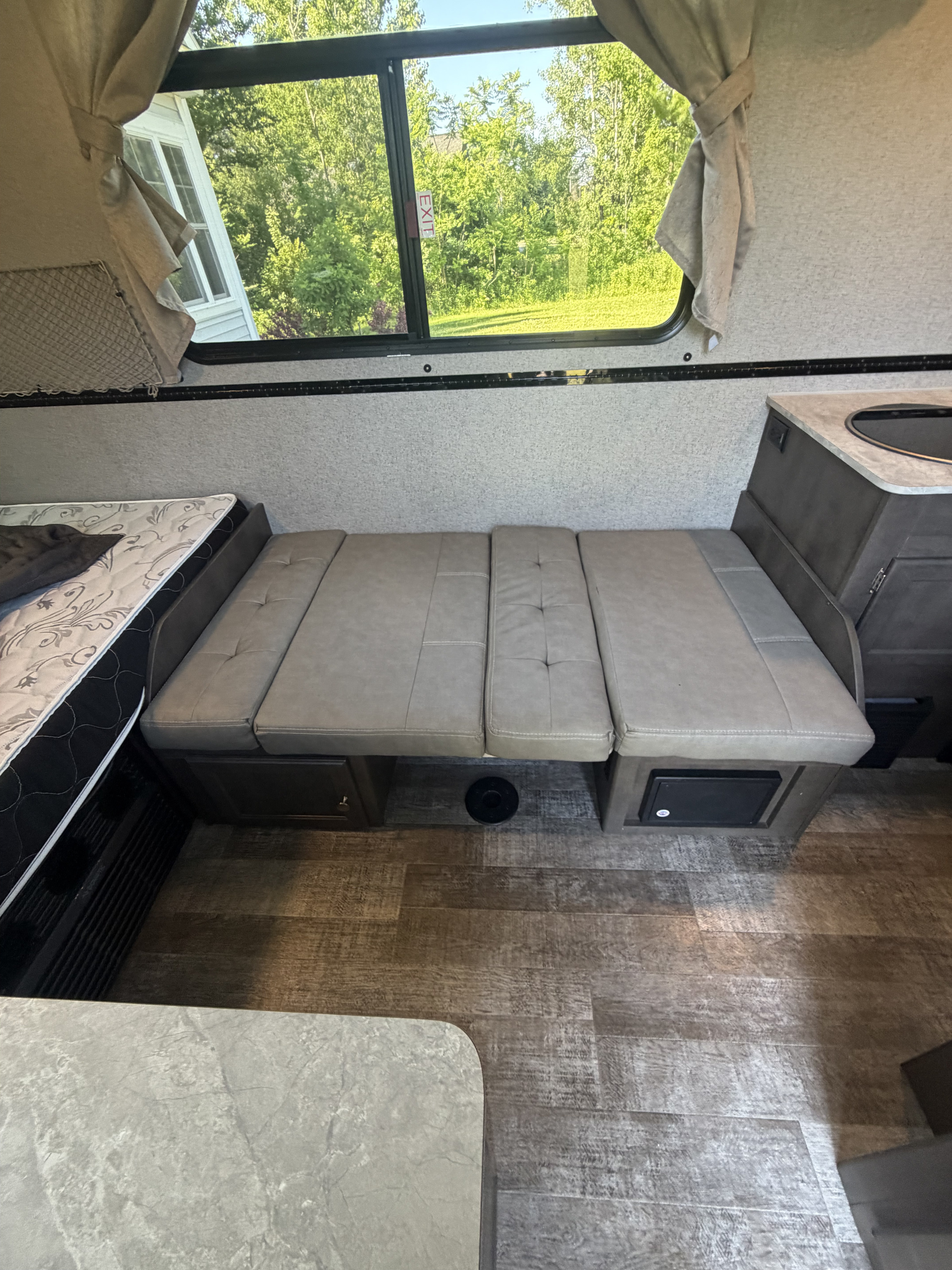Cozy RV rental interior featuring a convertible dinette, a bed, compact kitchen, and a window view of green trees.. Forest River Rockwood Premier 2021