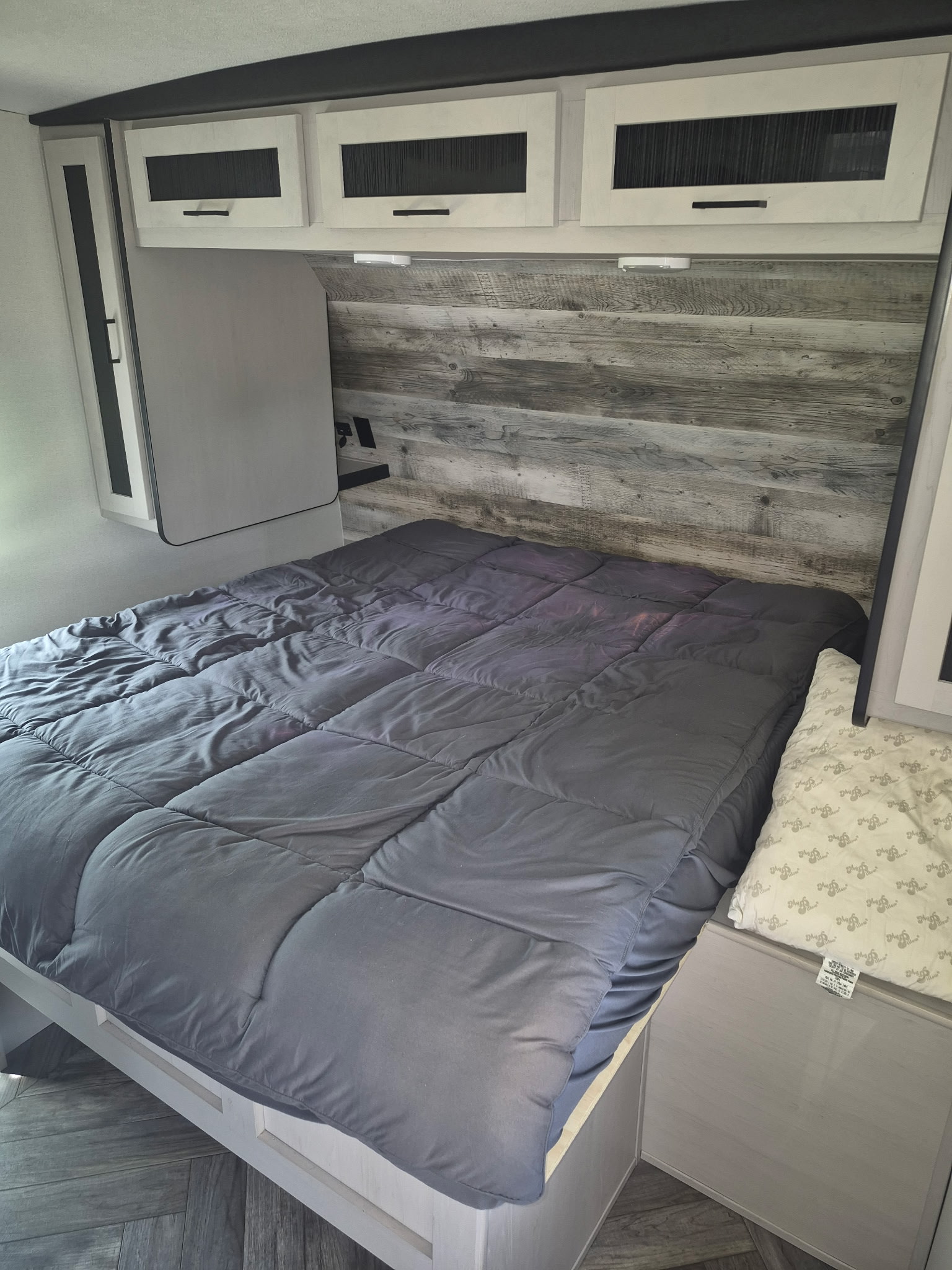 RV rental bedroom with a large bed, gray comforter, overhead storage, and rustic wood-paneled headboard.. Heartland RVs Mallard 2024