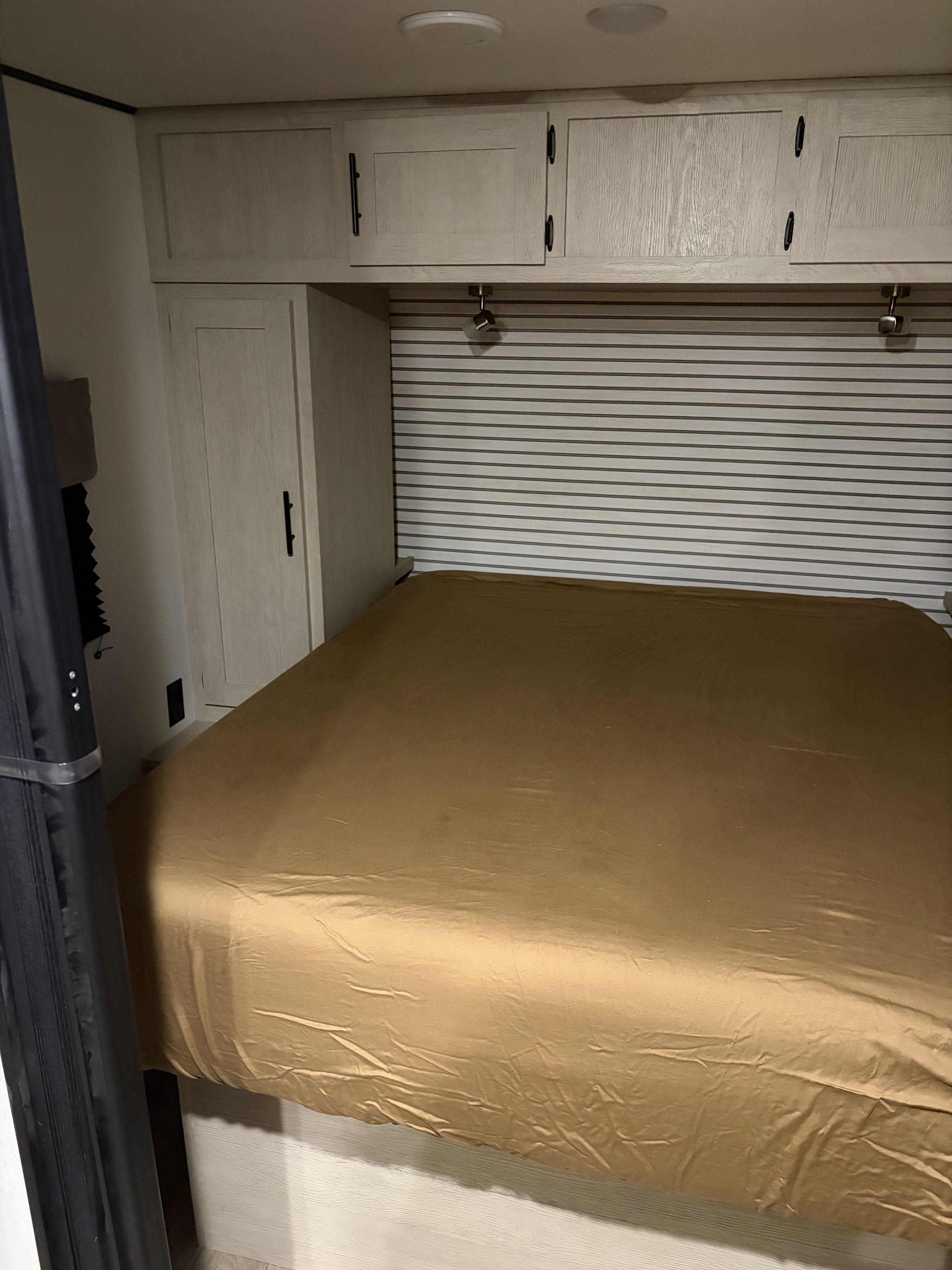 Inside an RV rental: a compact bedroom features a bed with golden-brown sheets, overhead cabinets, and reading lights.. Forest River Shasta I-5 2025