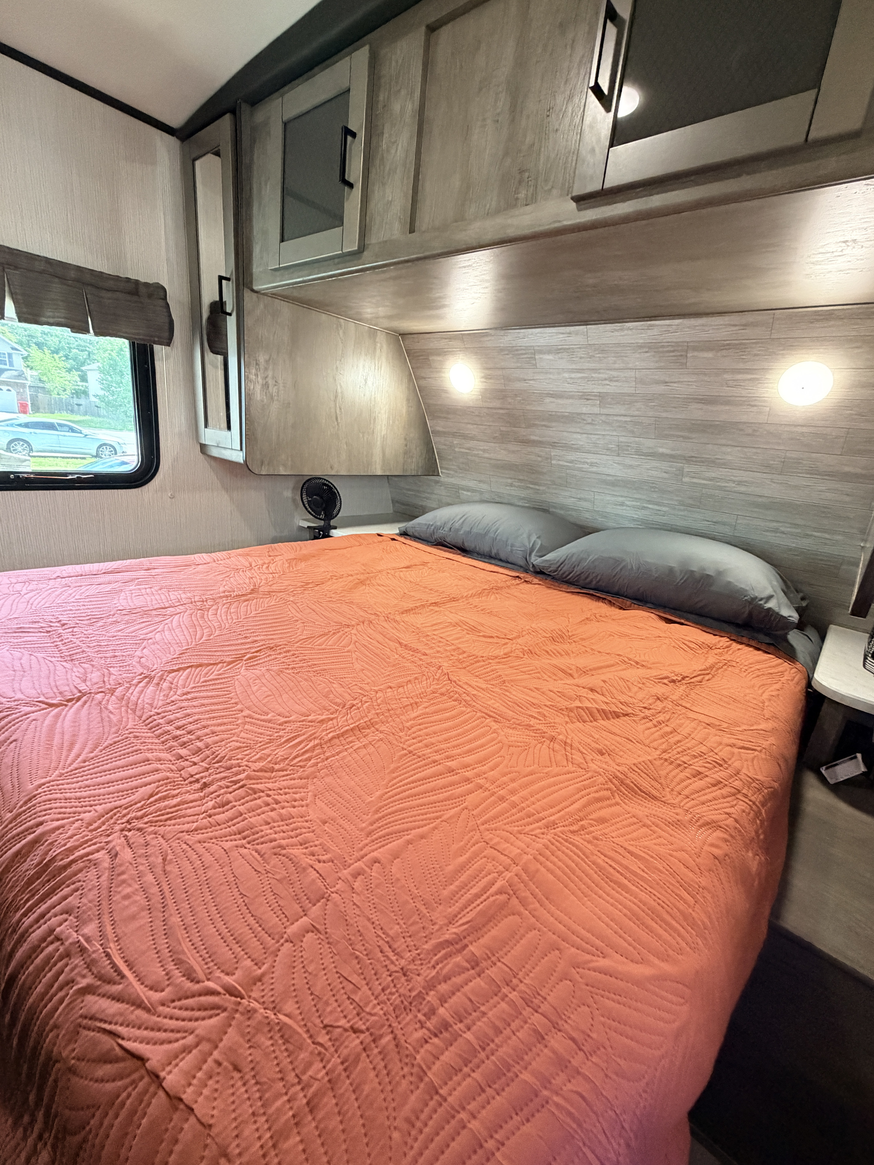 Cozy rv rental bedroom featuring a quilted orange bed, gray pillows, overhead storage, and a window.. Heartland RVs North Trail 2020