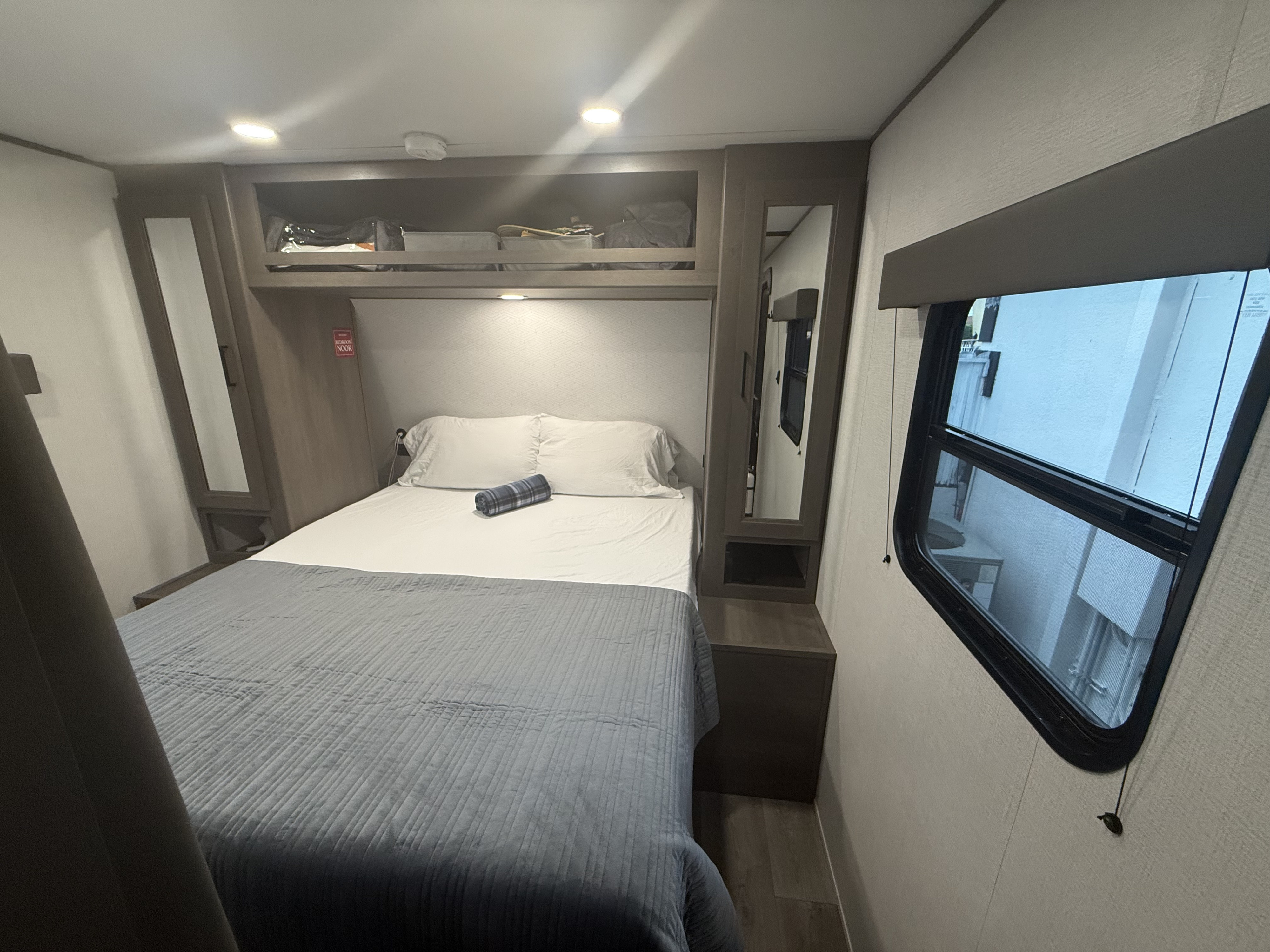 This RV rental bedroom offers a comfortable bed, overhead storage, a mirror, and a window for natural light.. Grand Design Transcend Xplor 2026