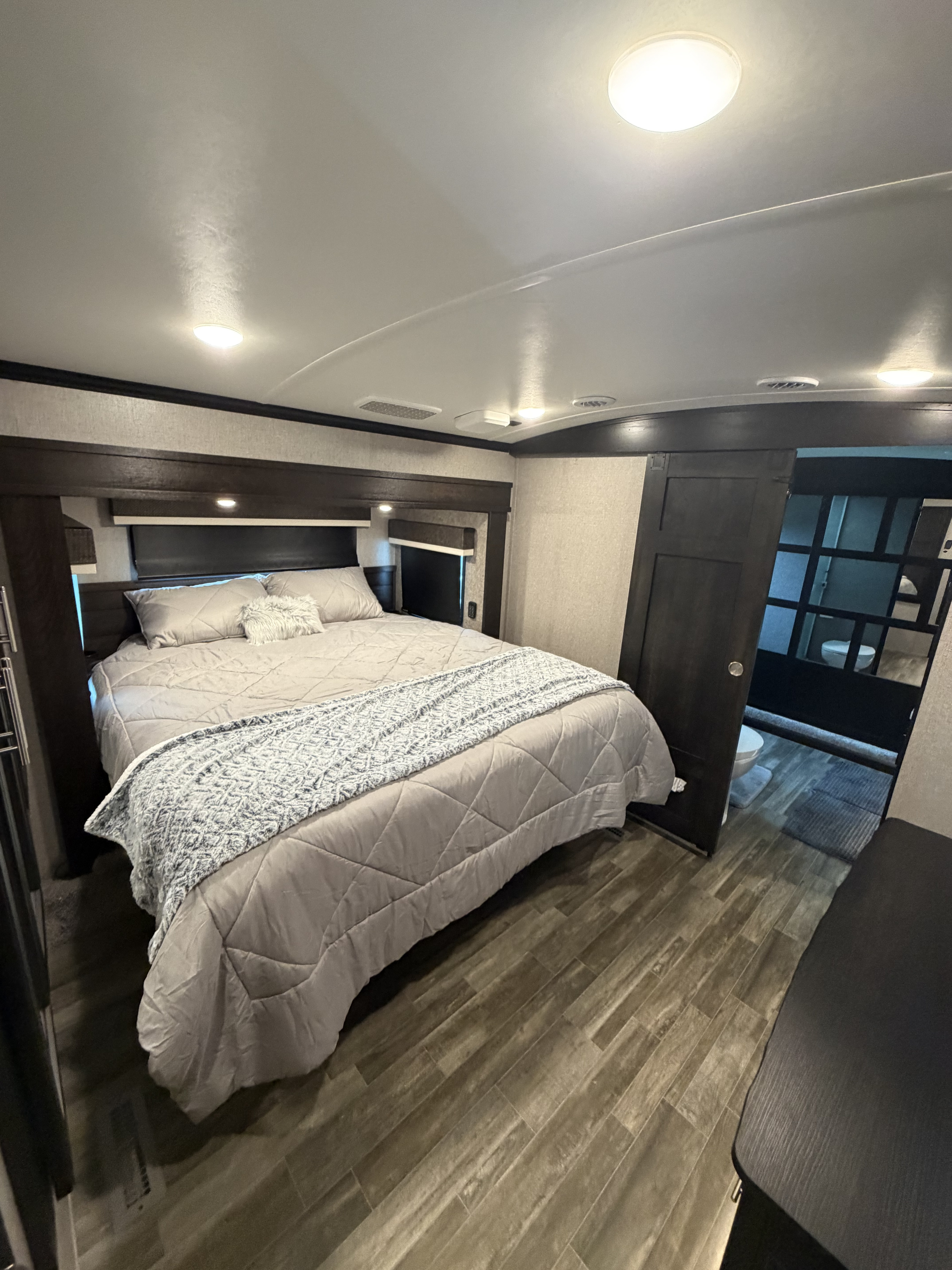 Cozy RV rental bedroom features a comfy gray bed, wood-look floor, and bathroom access. Brightly lit by ceiling lights.. Jayco North Point 2021