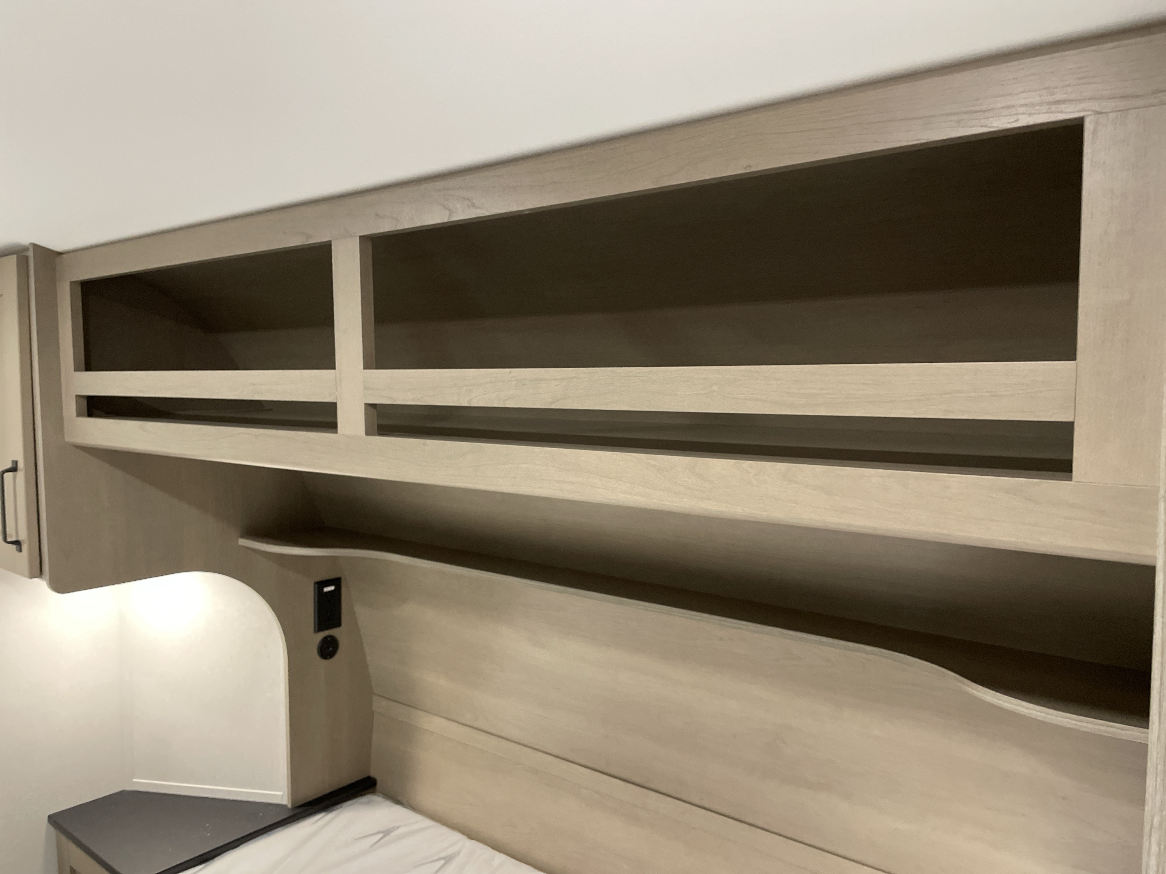 Cozy RV rental bedroom with light wood overhead storage, open shelving, and convenient bedside charging.. Grand Design Reflection 100 2026