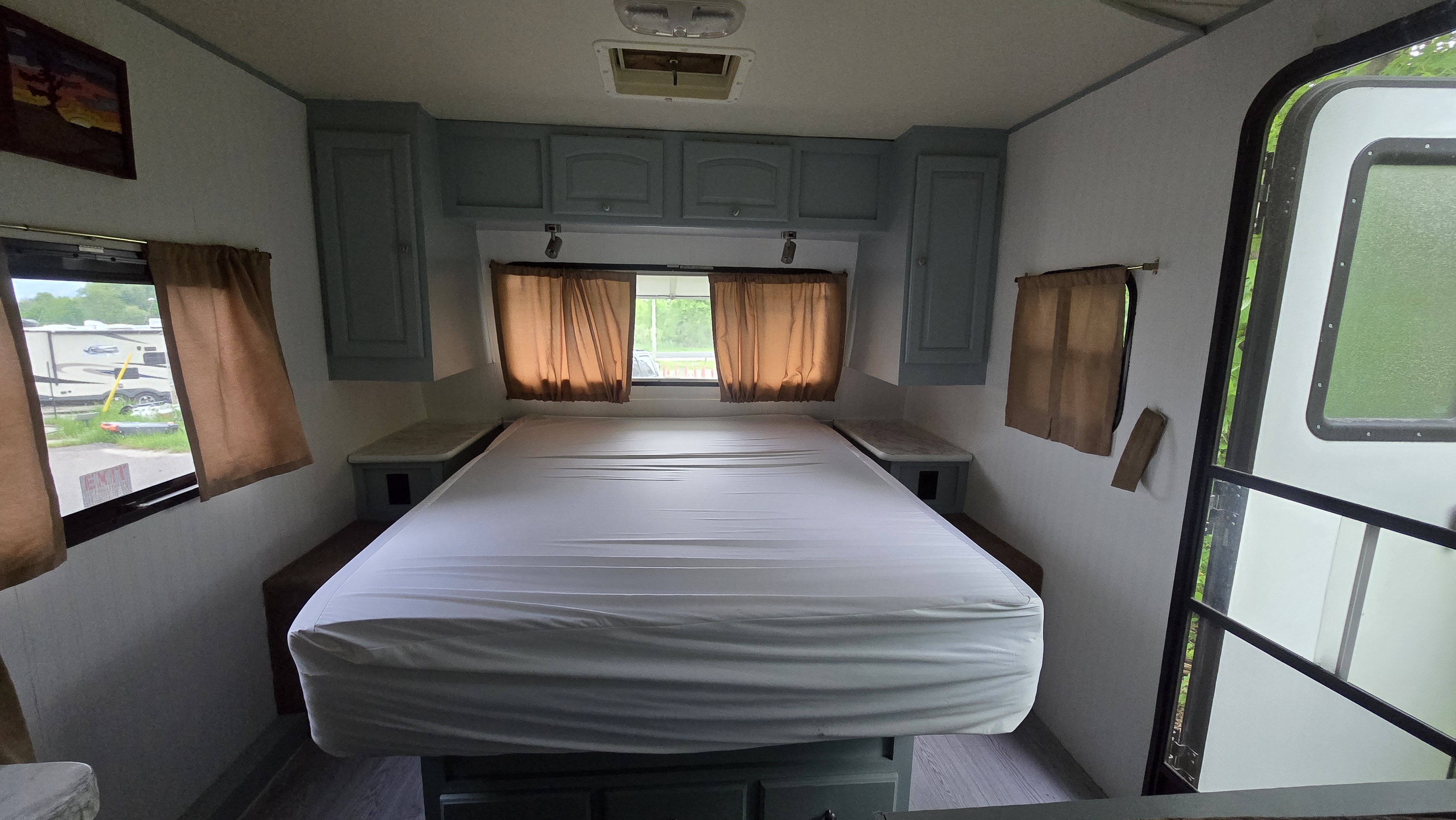 Clean and bright RV rental bedroom showing a large bed, blue storage cabinets, windows with brown curtains, and a side entrance.. Forest River Rockwood Mini Lite 2012